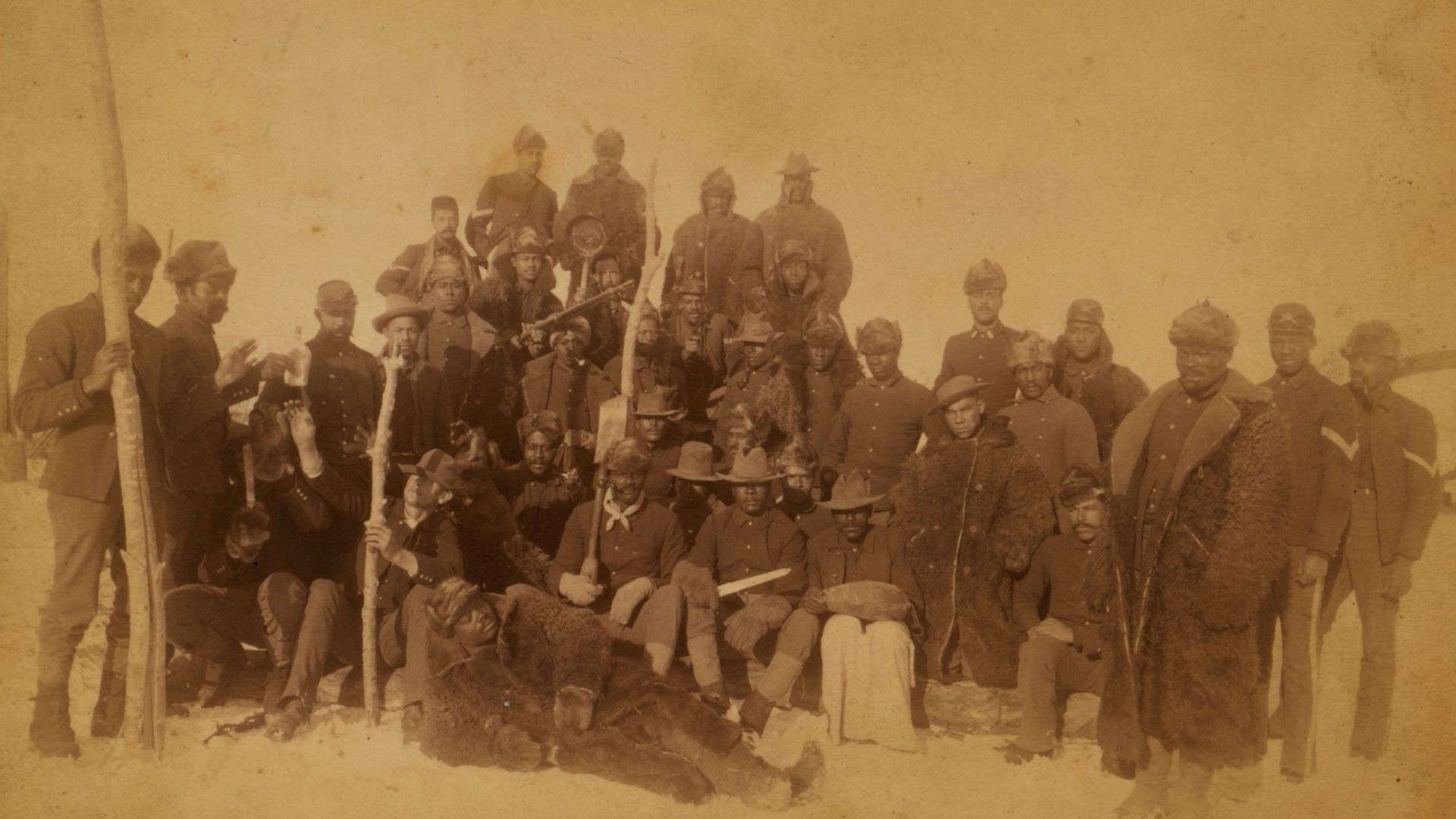 Sepia-toned group photo of soldiers in winter uniforms and fur coats, some holding staffs and rifles, posing outdoors on snowy ground in an old historic setting.