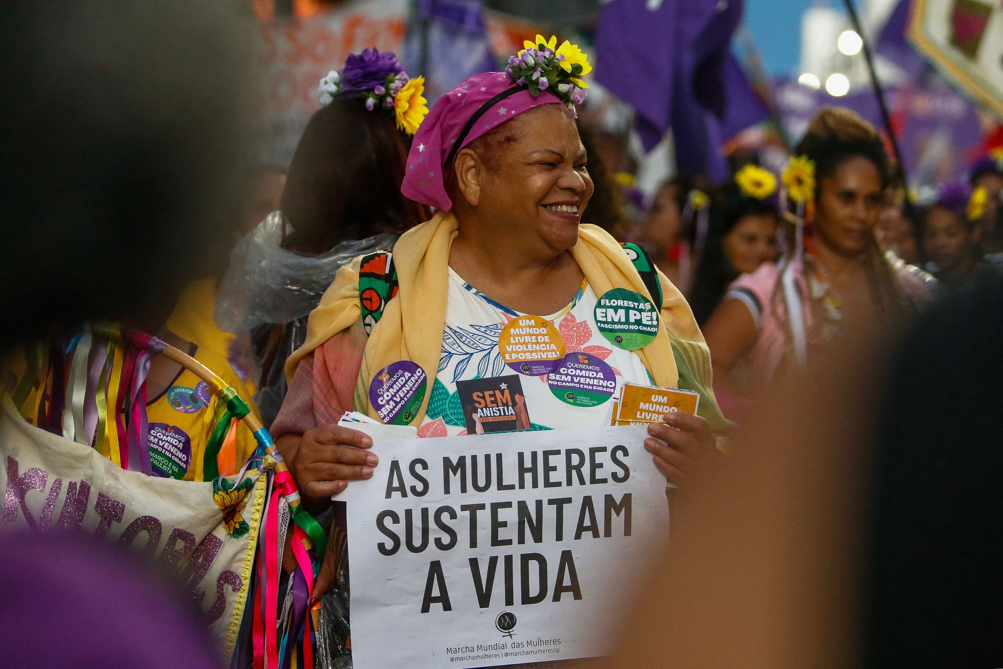a woman smiles and looks to the side while holding a sign in Portuguese that says "women sustain life"