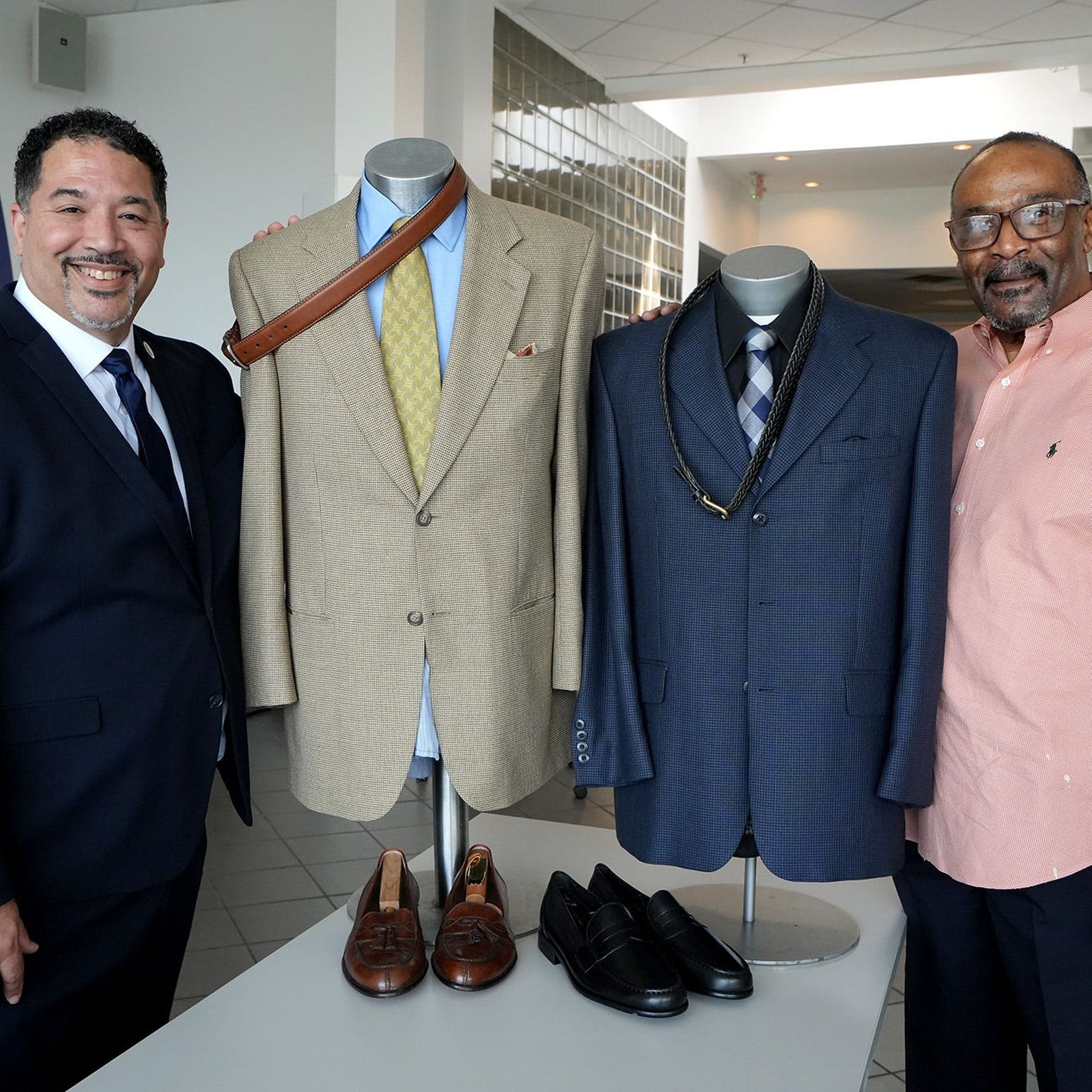 Two men stand beside suit displays at a nonprofit offering low-income men with professional warddrobes. 