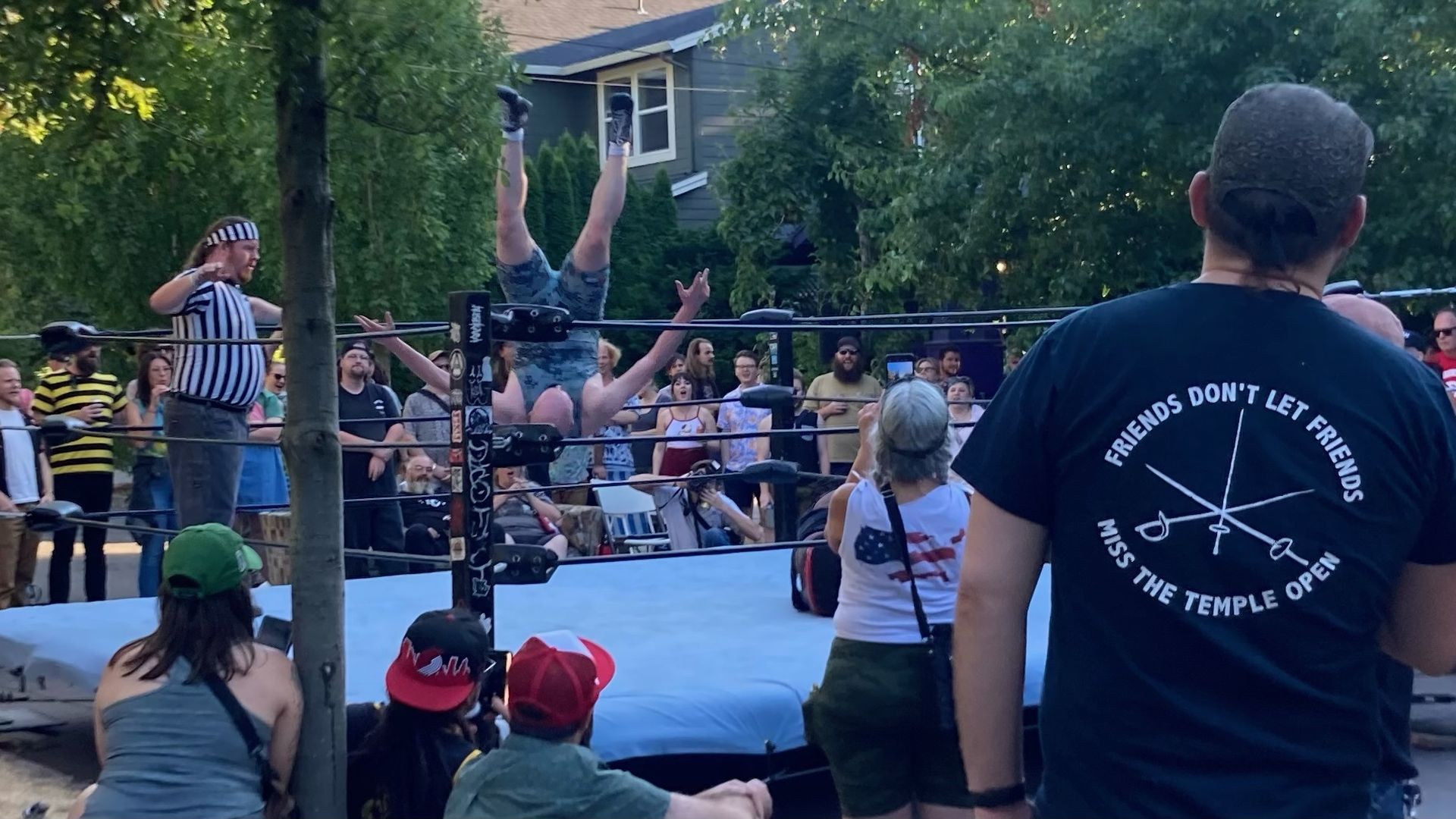 A man is upside down in the air, feet above his head, while a man in a black and white referee shirt looks on in a traveling wrestling ring set up with street onlookers.