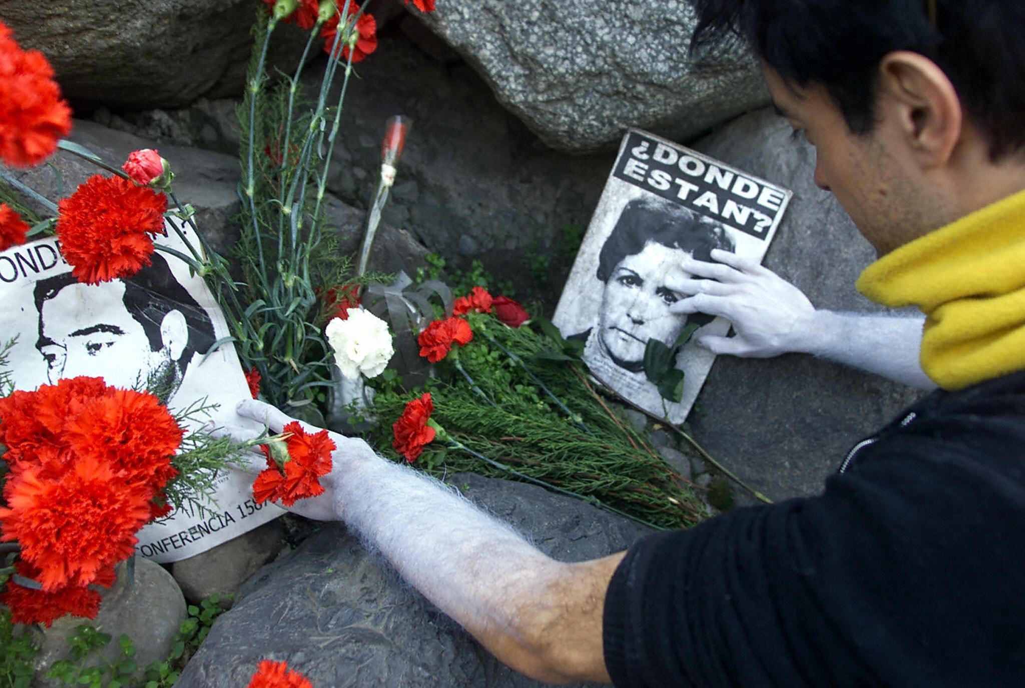 a man with white paint covering his hands and forearms places red flowers on the ground next to a photo of a woman who went missing during the Augusto Pinochet regime in Chile