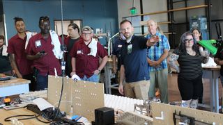 A man points to a table full of electronic equipment while leading a tour group