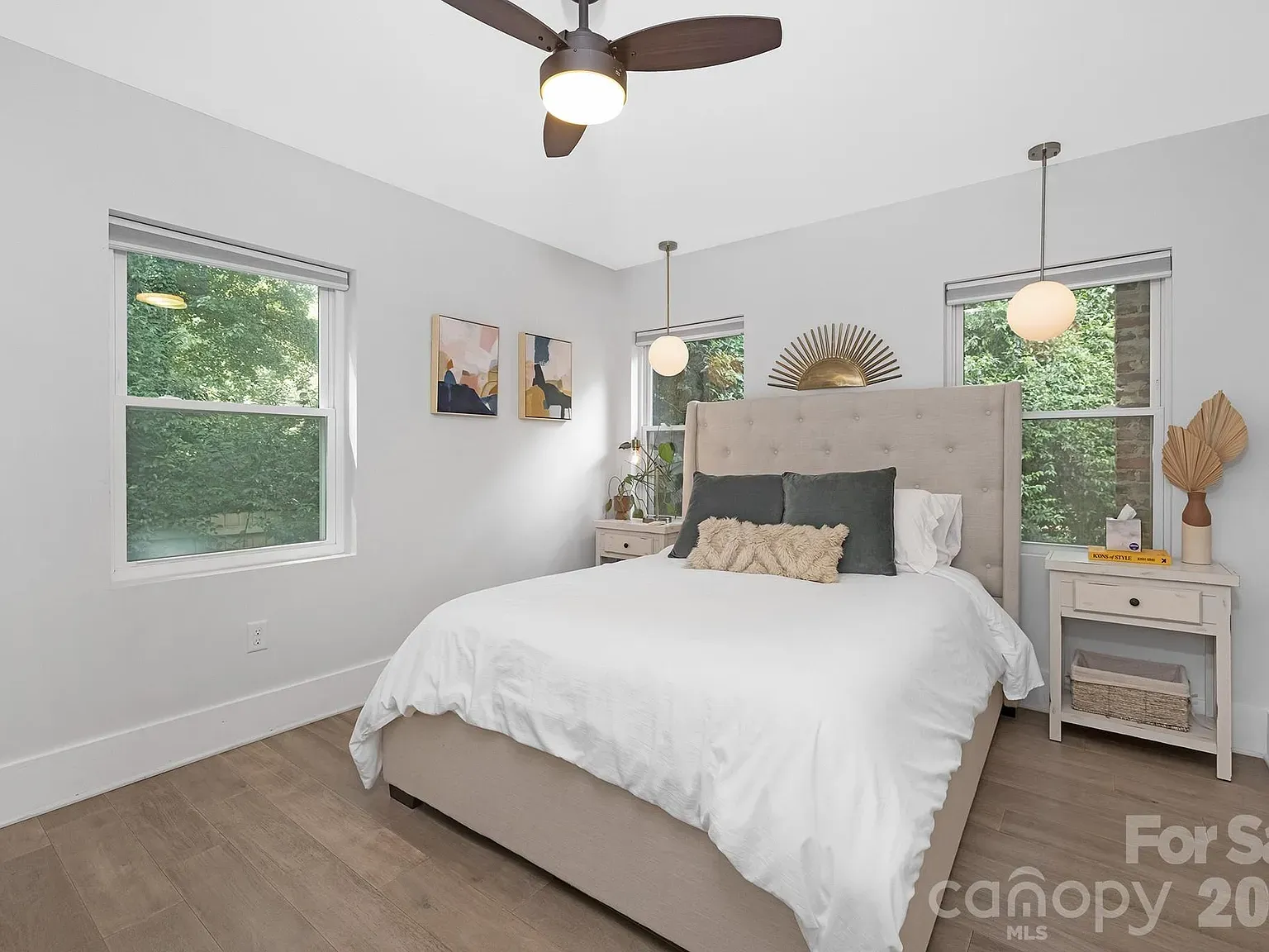 Bright modern bedroom with a beige upholstered bed, white bedding, two nightstands, round pendant lights, a ceiling fan, large windows with greenery outside, and abstract wall art.