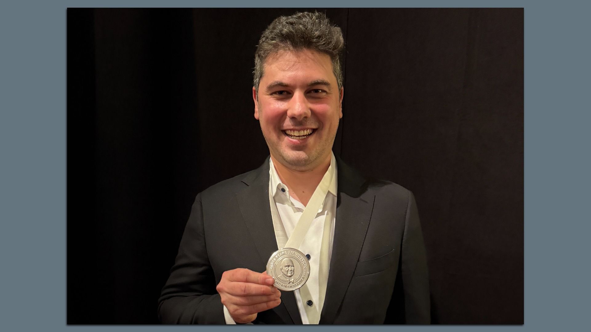 Smiling man in a dark suit and white shirt holds a large round silver medal on a white ribbon, posed against a dark backdrop.