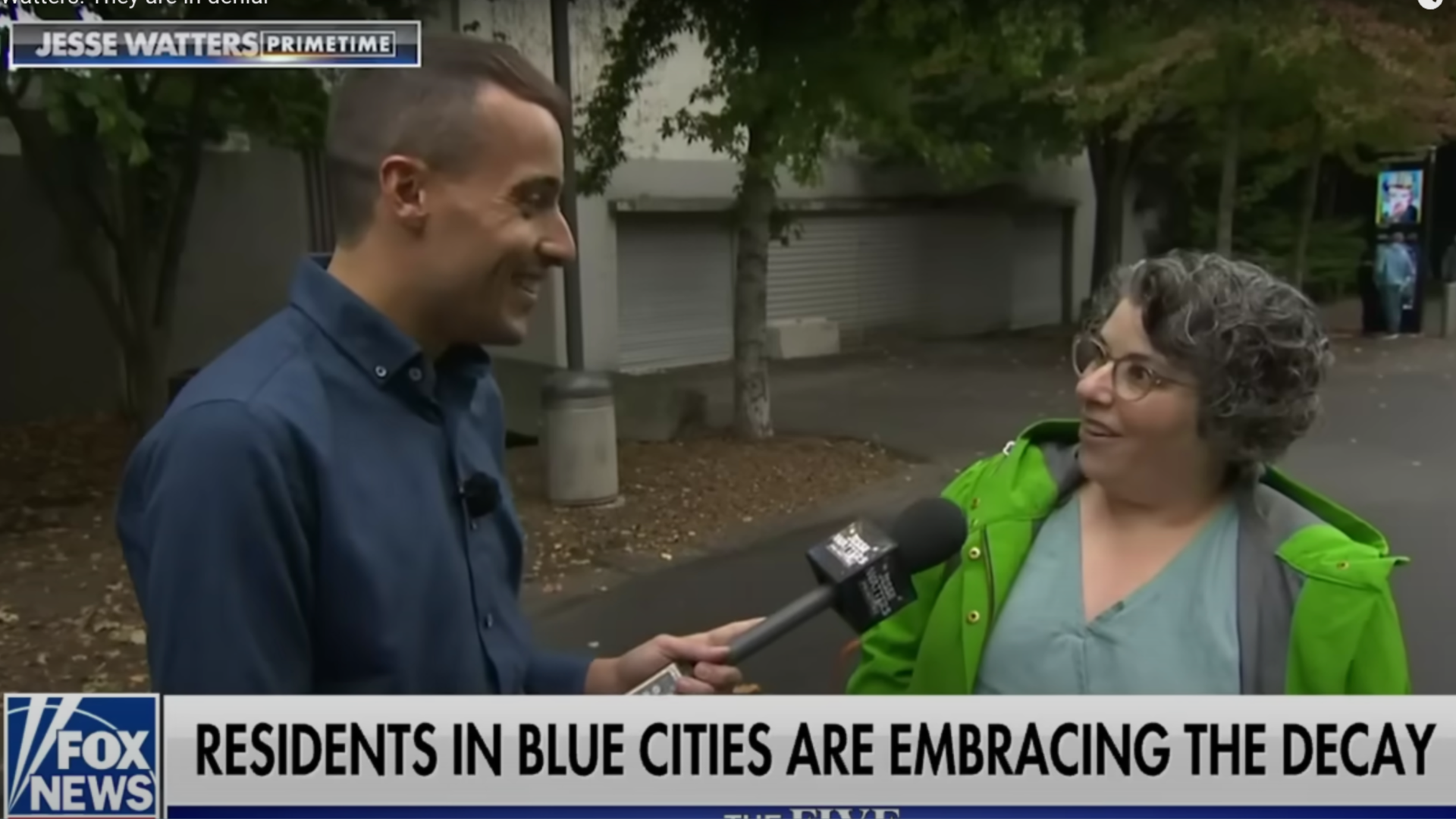 A Fox interviewer in a blue button up shirt holds a mic to a shorter person in a bright green jacket as she speaks into the mic.