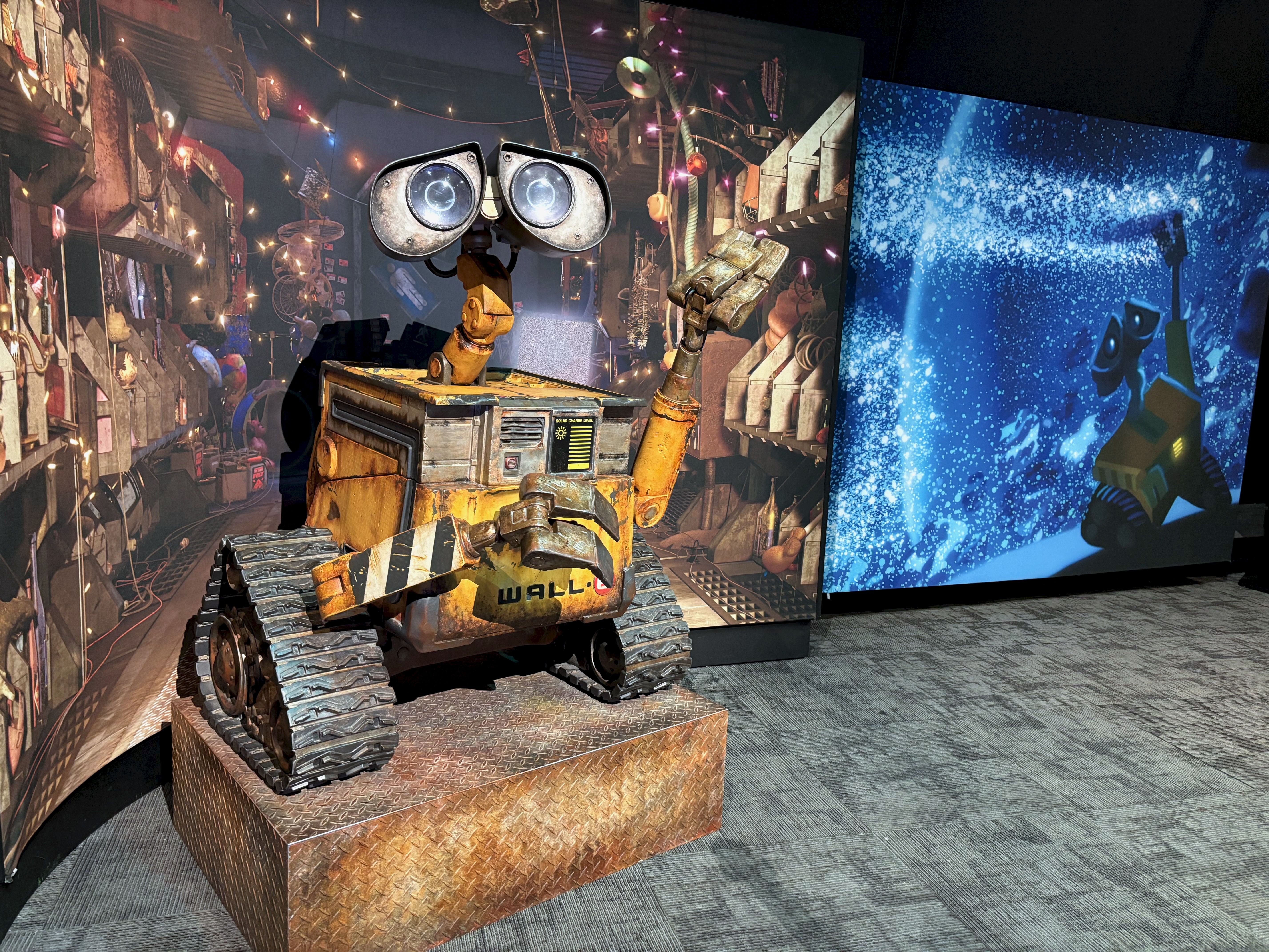Model of the yellow robot WALL-E on a metal platform in front of a detailed workshop backdrop and adjacent screen showing WALL-E in a starry blue scene from the animated movie.