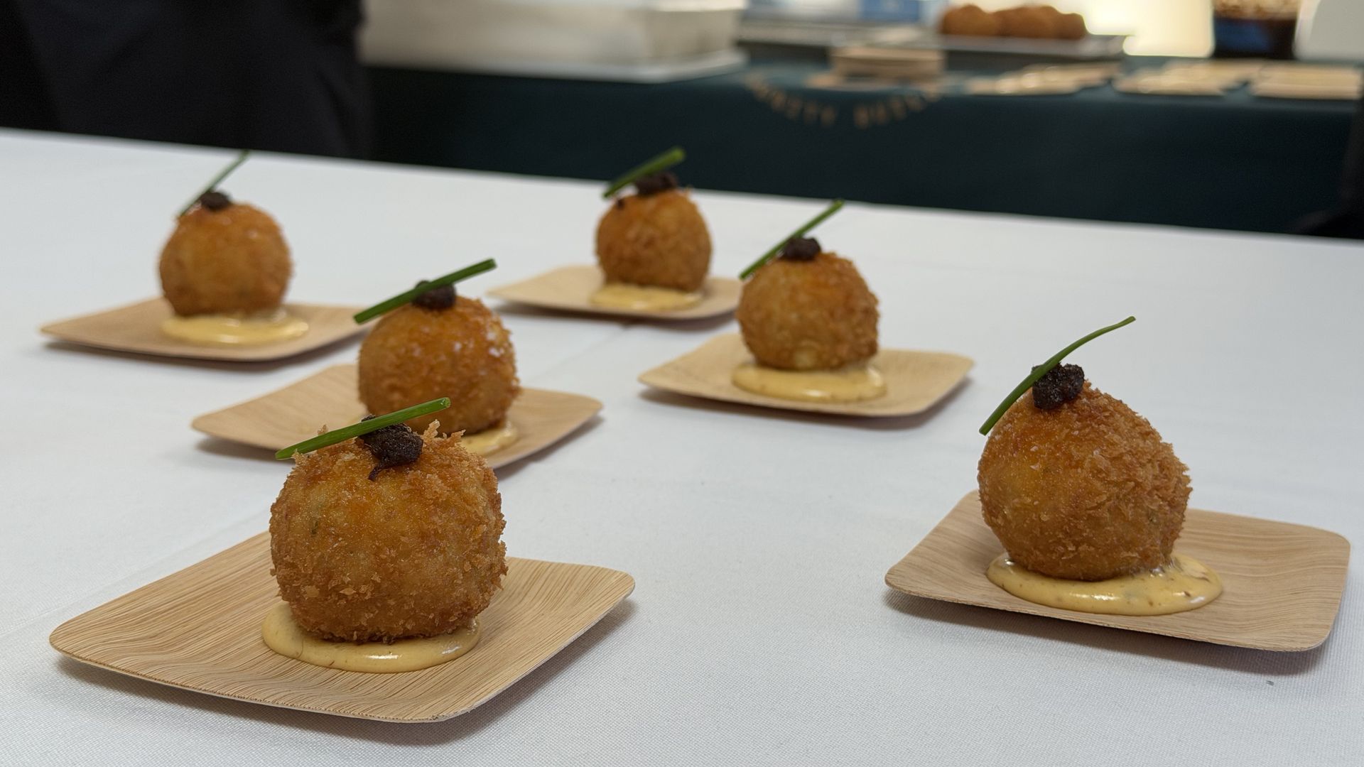 Row of golden fried croquettes made with flounder and cassava sit on shito sauce, placed on wooden plates at a white table that's Comfort Kitchen's station for a flounder event.