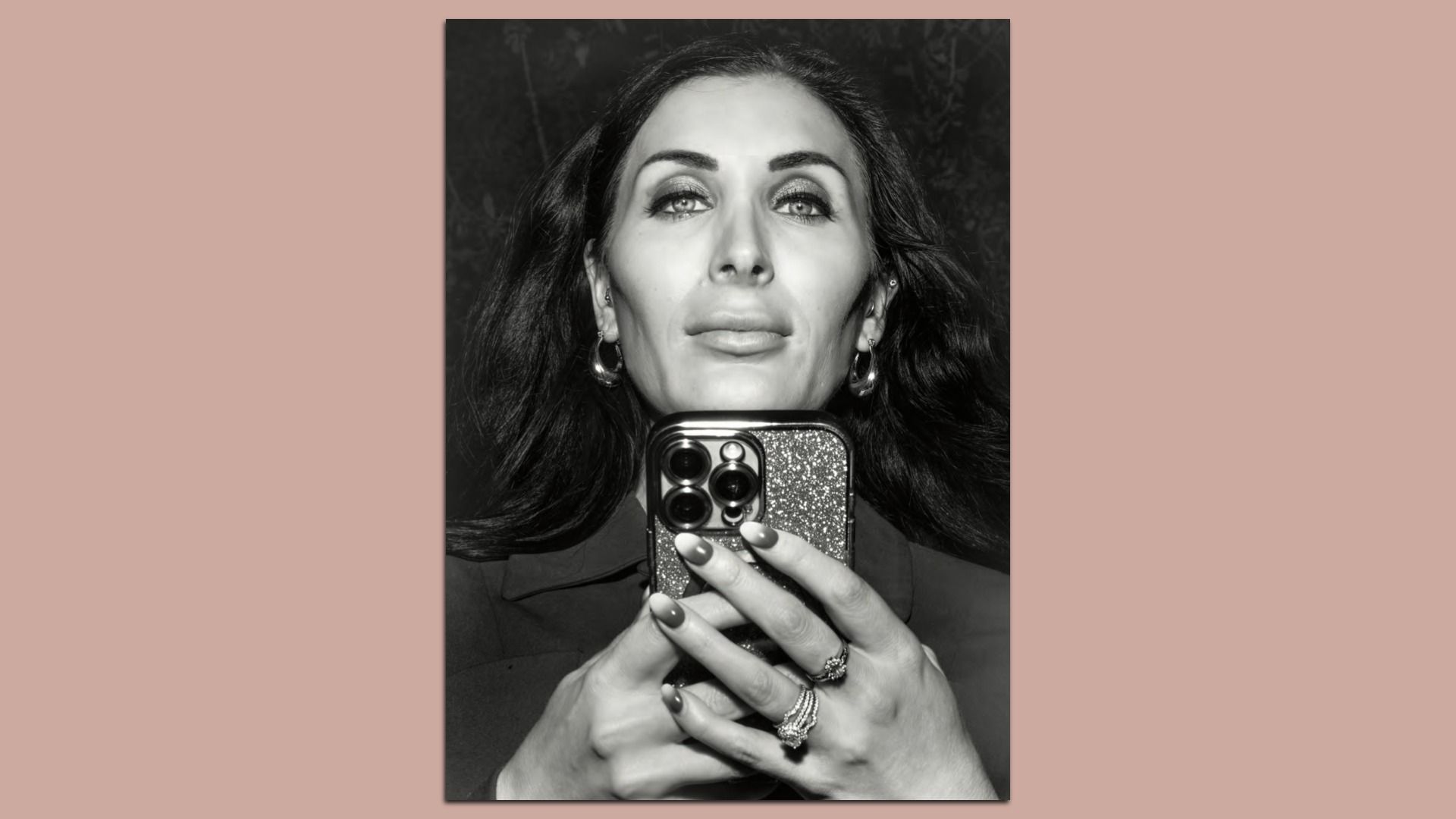 Black and white photo of a woman with long dark hair holding a phone with a glittery case, wearing hoop earrings, rings, and a collared shirt, looking confidently at the camera.