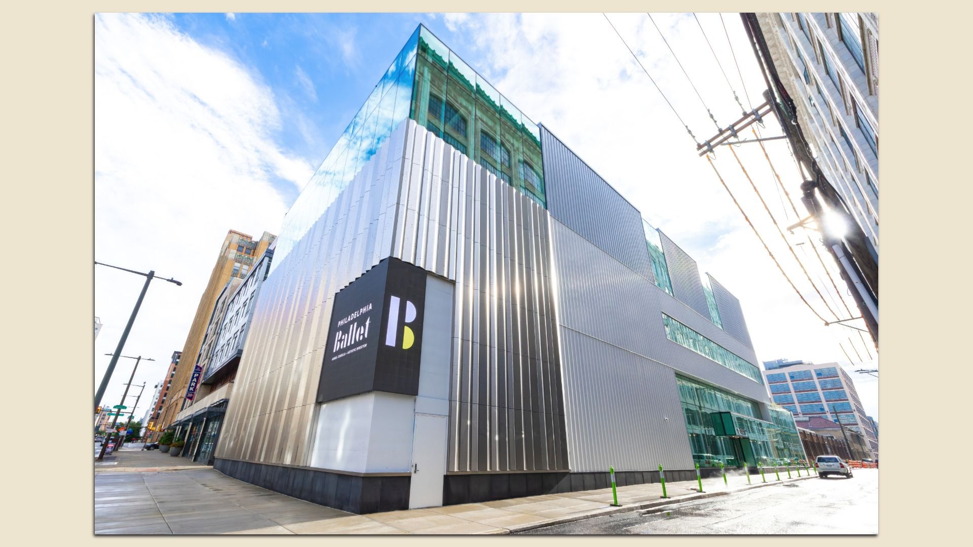 Modern building clad in vertical corrugated metal panels with a glass upper section on a city street; blue sky, overhead power lines, and a sign reading "Philadelphia Ballet".