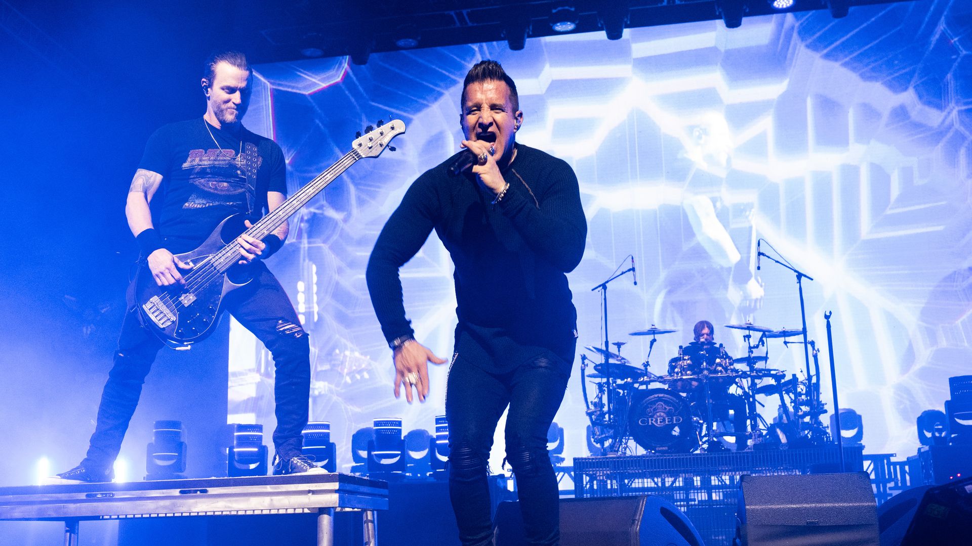 The band Creed performs on stage, with lead singer Scott Stapp and a guitarist facing the camera and a patterned lighted backdrop is shown behind the stage.