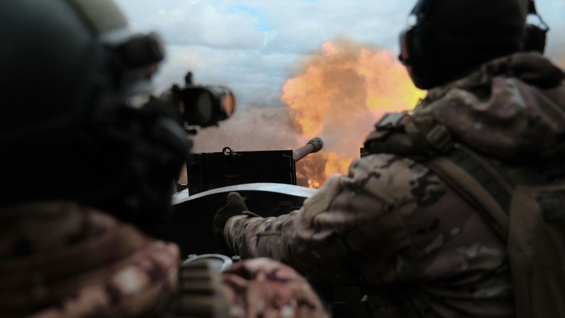 A large artillery piece fires. An orange fireball exits the barrel. Two men are on either side of it. The sky is blue.