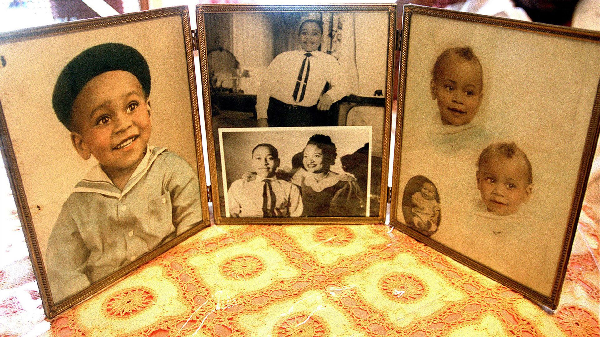 Three framed vintage photographs on a yellow and orange lace tablecloth: a smiling Emmett Till as a boy in a sailor outfit and beret, two black-and-white portraits of a boy in a tie and a woman, and two baby portraits.