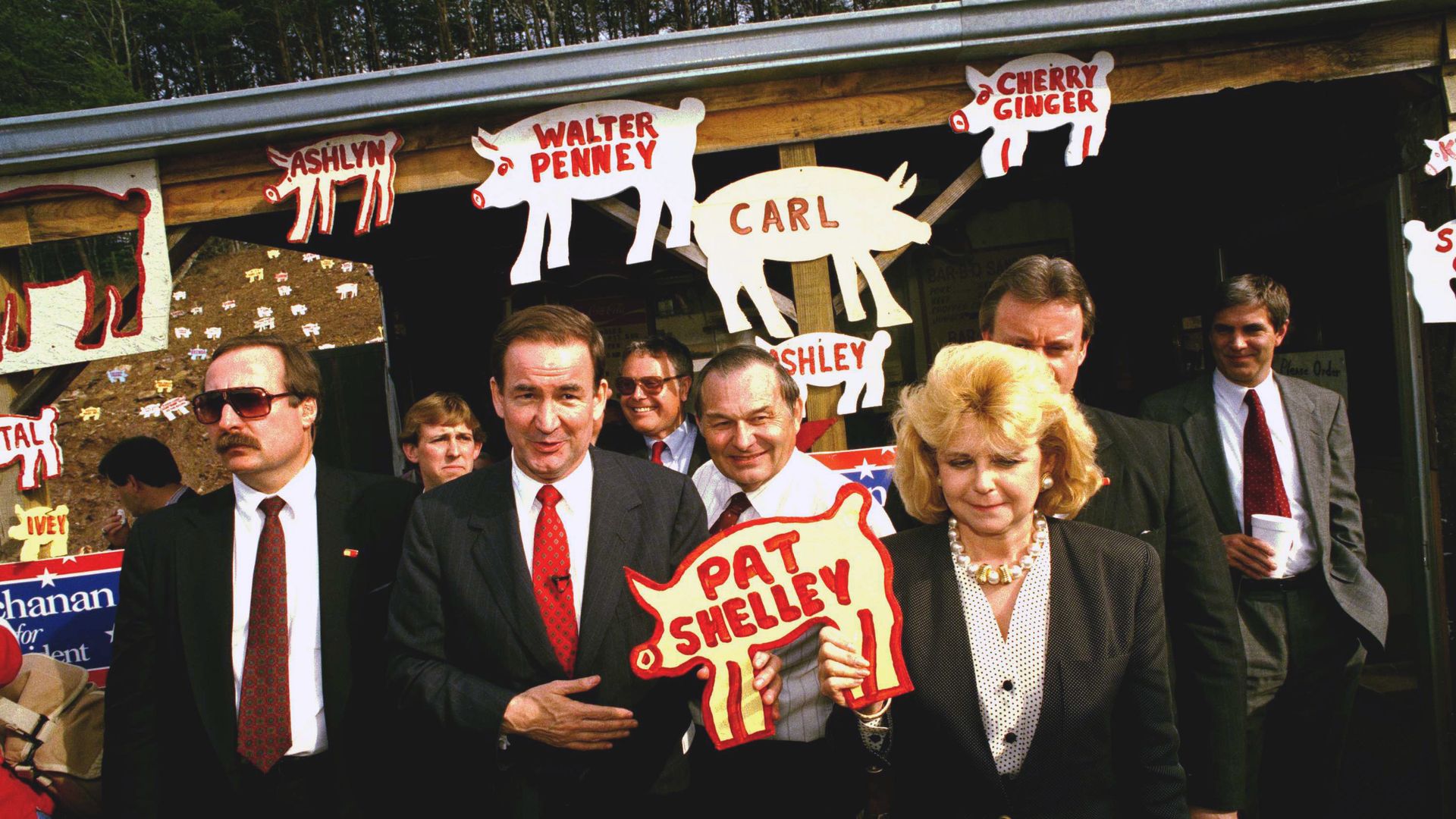 A photo of Pat Buchanan posing with a wooden pig with his and his wife Shelley's names in front of a barbecue restaurant