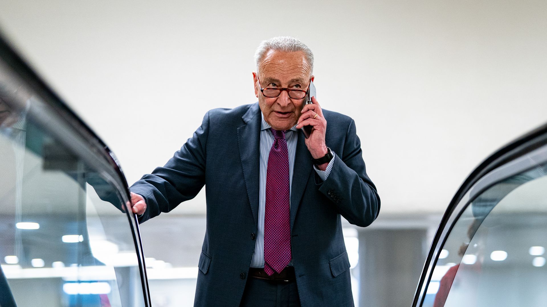 An older man in a dark blue suit, purple tie, and glasses is talking on a cellphone while standing on an escalator in a modern, well-lit indoor space.