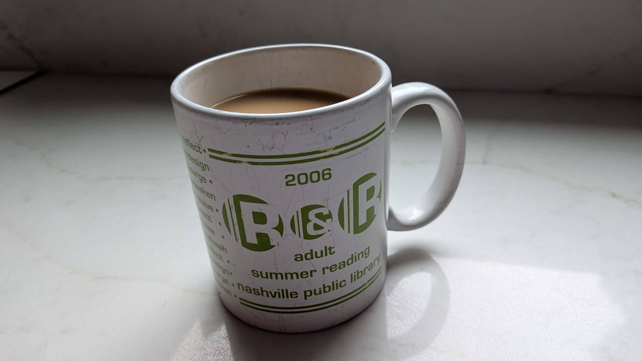 White ceramic mug half-full of light brown coffee sits on a pale marble surface. It has green printed text and lines, including "2006", with a worn, scratched exterior.