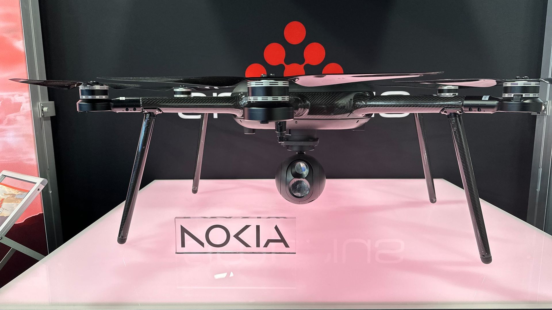 Black quadcopter drone with four propellers and a spherical camera mounted underneath, displayed on a white surface with a red logo and the word "NOKIA" visible in the background.