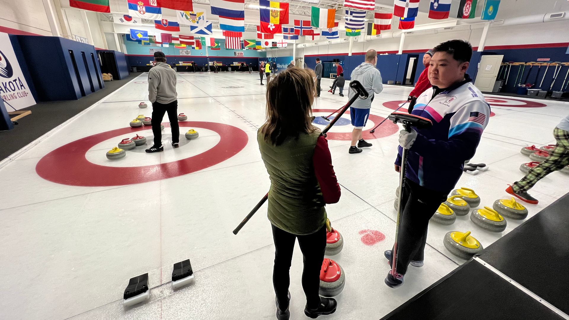 Two players stand on a curling rink — a white sheet of ice with bullseye-shaped targets of red rings, with special grey curling stones with yellow and red tops. The players are in conversation and holding curling brooms, which have flat heads and no bristles.