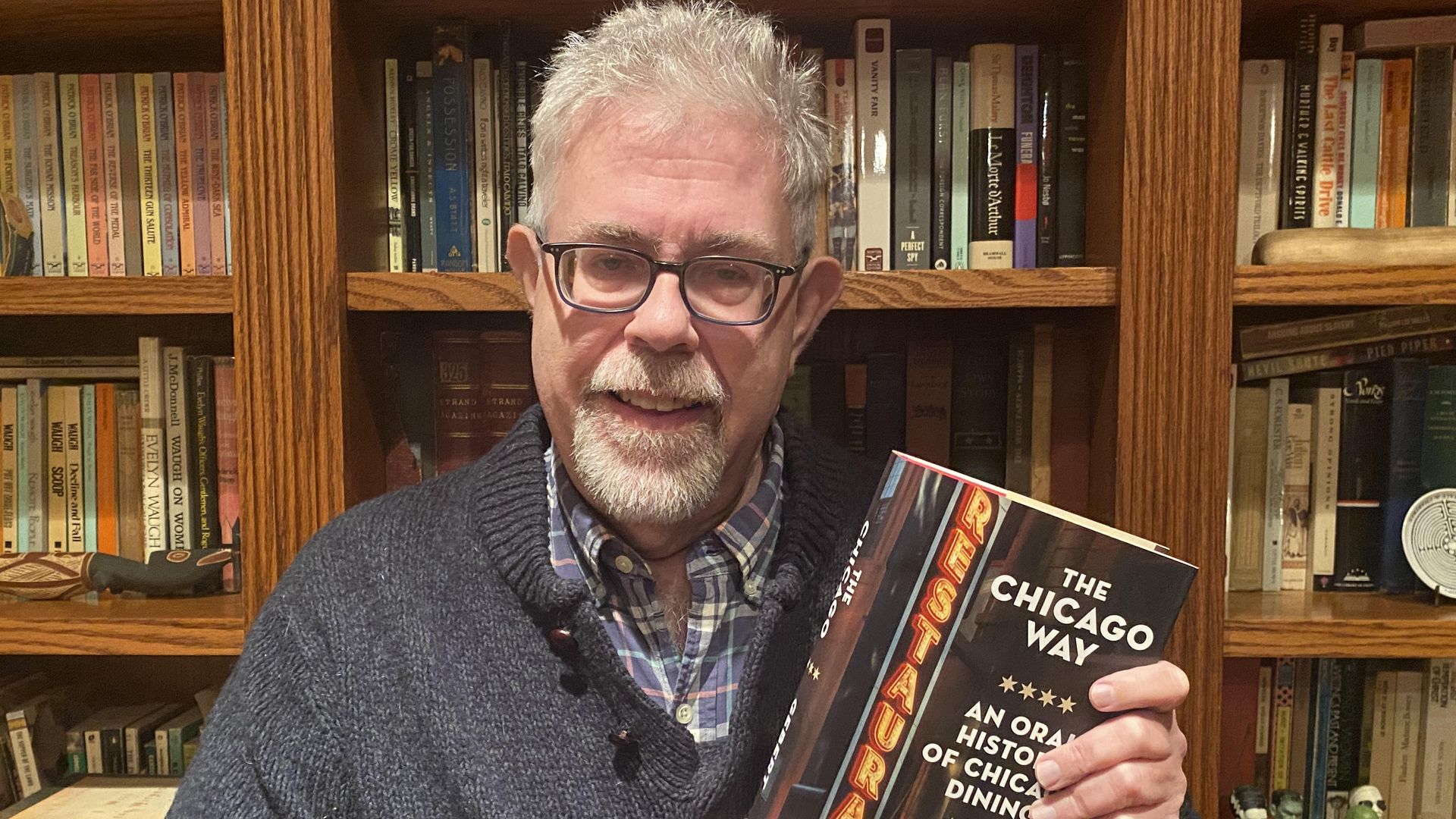 An older man with glasses, gray hair, and beard wearing a navy sweater and plaid shirt holds the book "The Chicago Way: An Oral History of Chicago Dining" by Michael Gebert in front of a wooden bookshelf.
