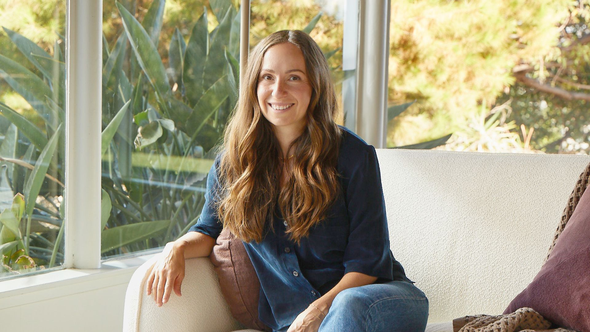Ariel Kaye, the CEO and founder of Parachute, sits on a white sofa in a living room with floor to ceiling windows.