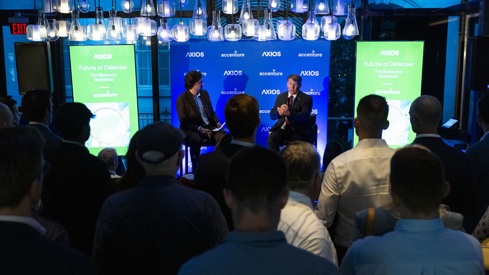 Two men in suits sit in bar-height chairs and are holding mics. Lots of people are seen in the foreground, blurred out of focus.