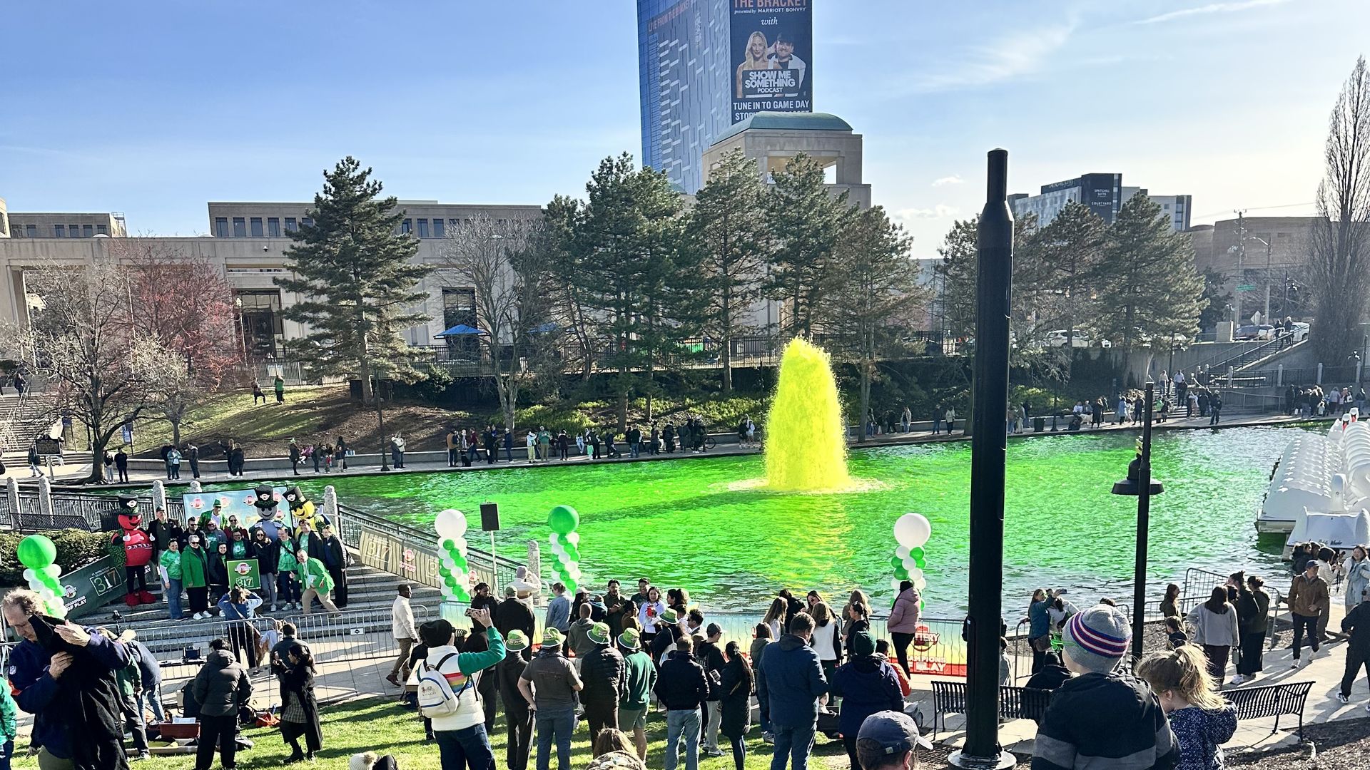 Sunny city park with a green fountain in a rectangular pond, crowds on the grass and along the railing; people wear green as balloons line the walkway and a tall building rises in the background.