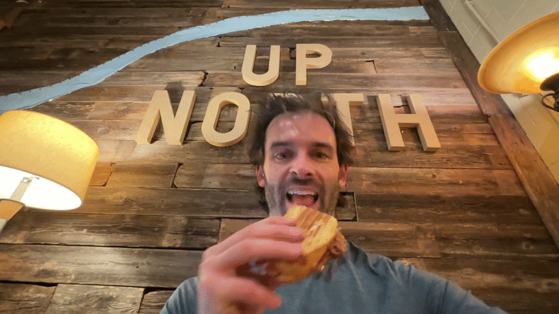 Gif shows reporter Travis biting into a sandwich with "UP NORTH" signage behind him and lamps on either side.