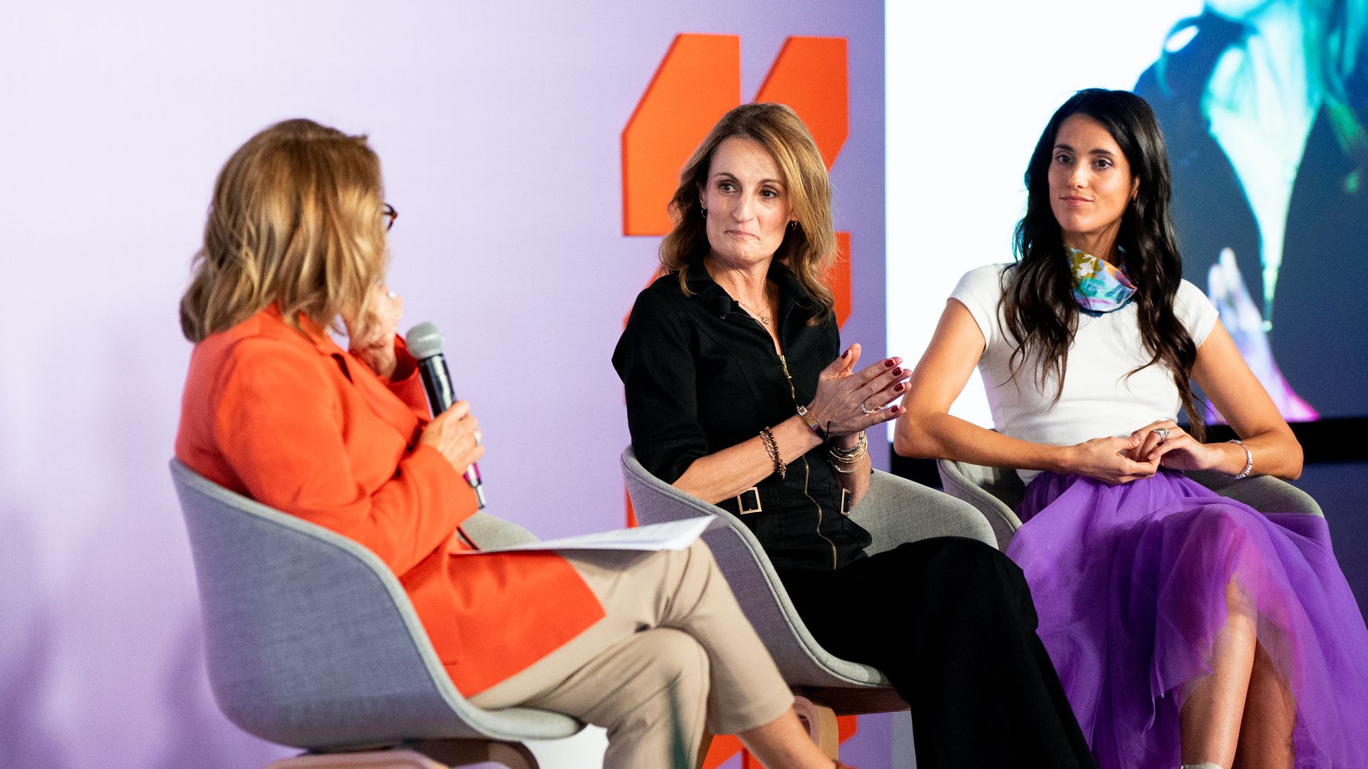 Three people on a panel stage: left moderator in an orange jacket holds a microphone, middle in black, right in white top with a purple tulle skirt; lavender backdrop with orange shapes.