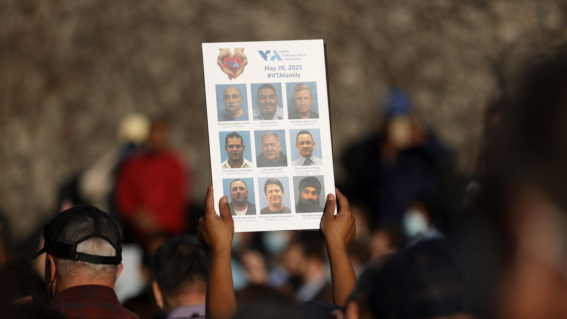 Photo of two hands holding up a sign with nine photos, one for each of the men killed by the San Jose mass shooter
