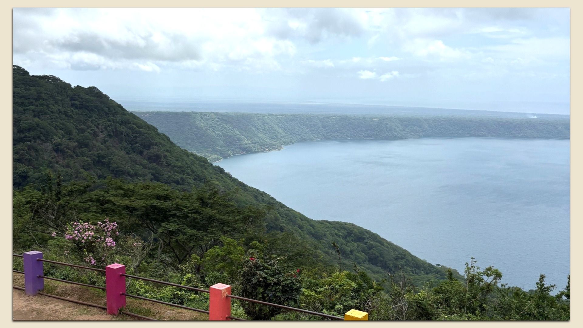 View from a hillside overlook of a large blue lake surrounded by green forested hills; a railing with pastel purple, pink, orange, and yellow posts in the foreground; cloudy sky above.