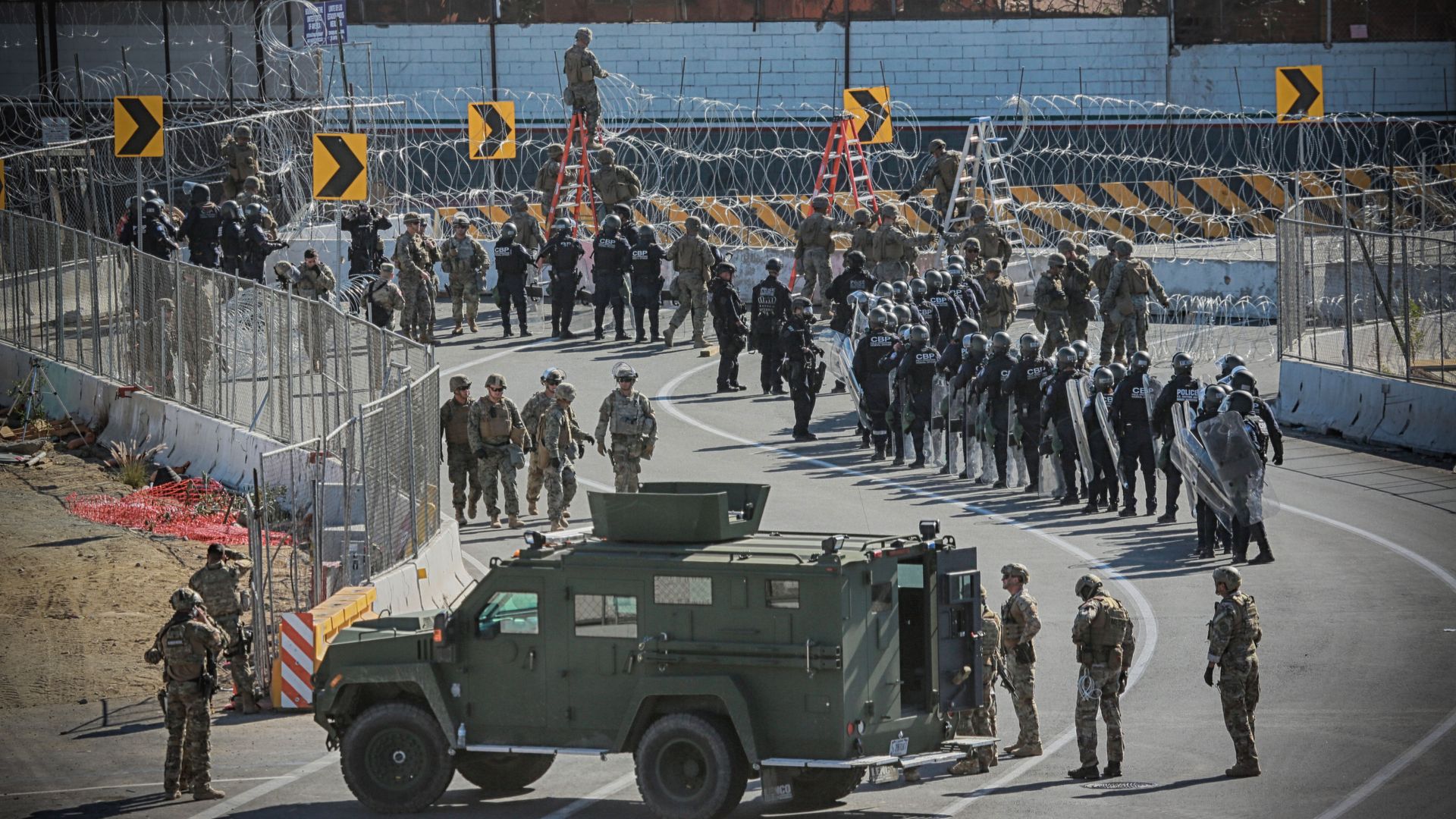 Troops and border patrol at the border where there is barbed-wire covered fence. 