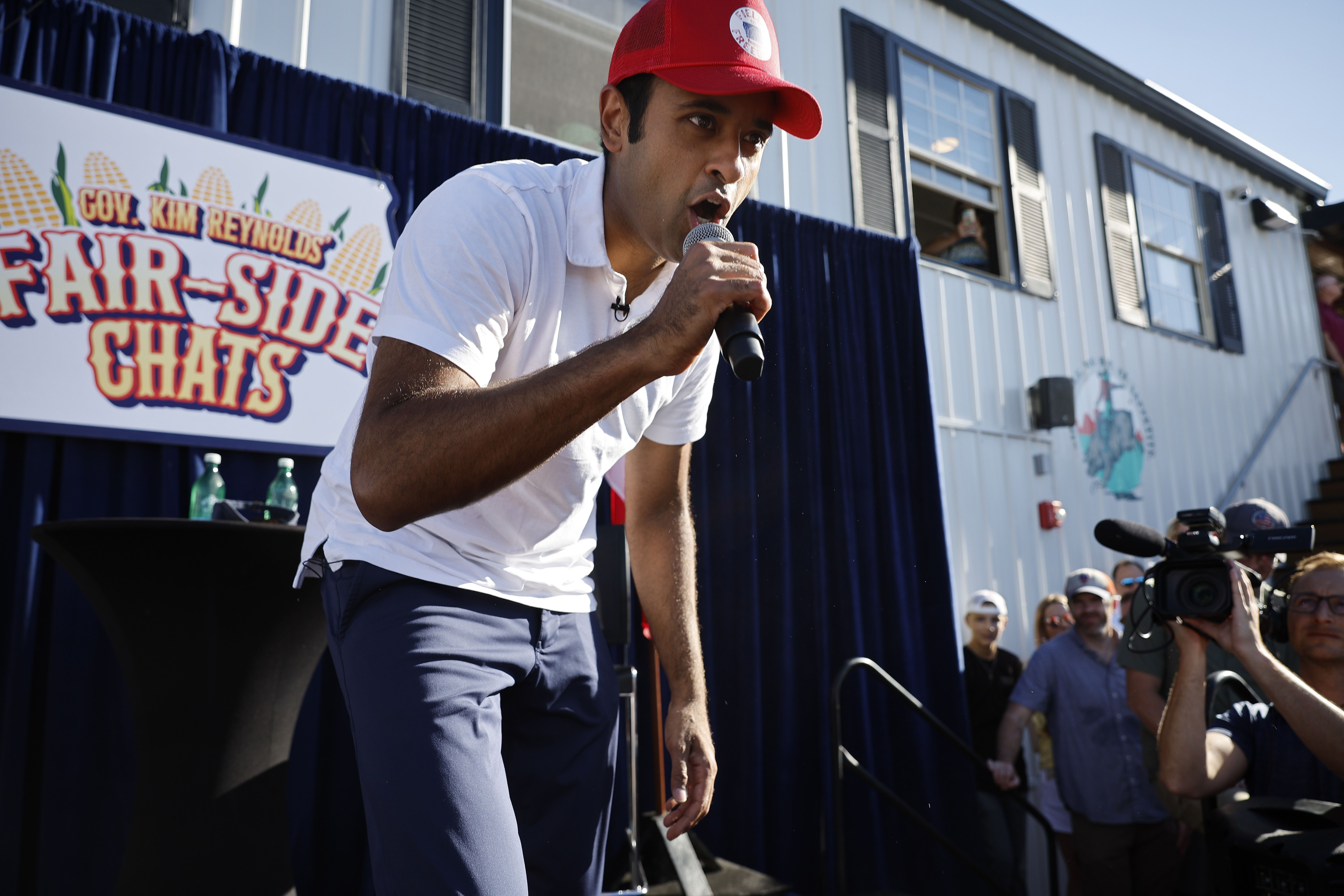 Vivek Ramaswamy speaks into a microphone on an Iowa State Fair stage. Behind him is a poster reading "Gov. Kim Reynolds' Fair-Side Chats."