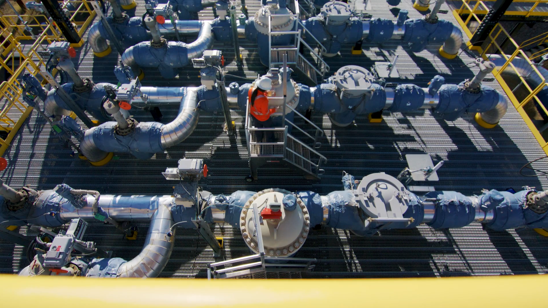 Overhead view of an industrial plant with blue pipes, valves and metal grating. A worker in an orange safety vest and white hard hat stands on a small platform inspecting equipment.