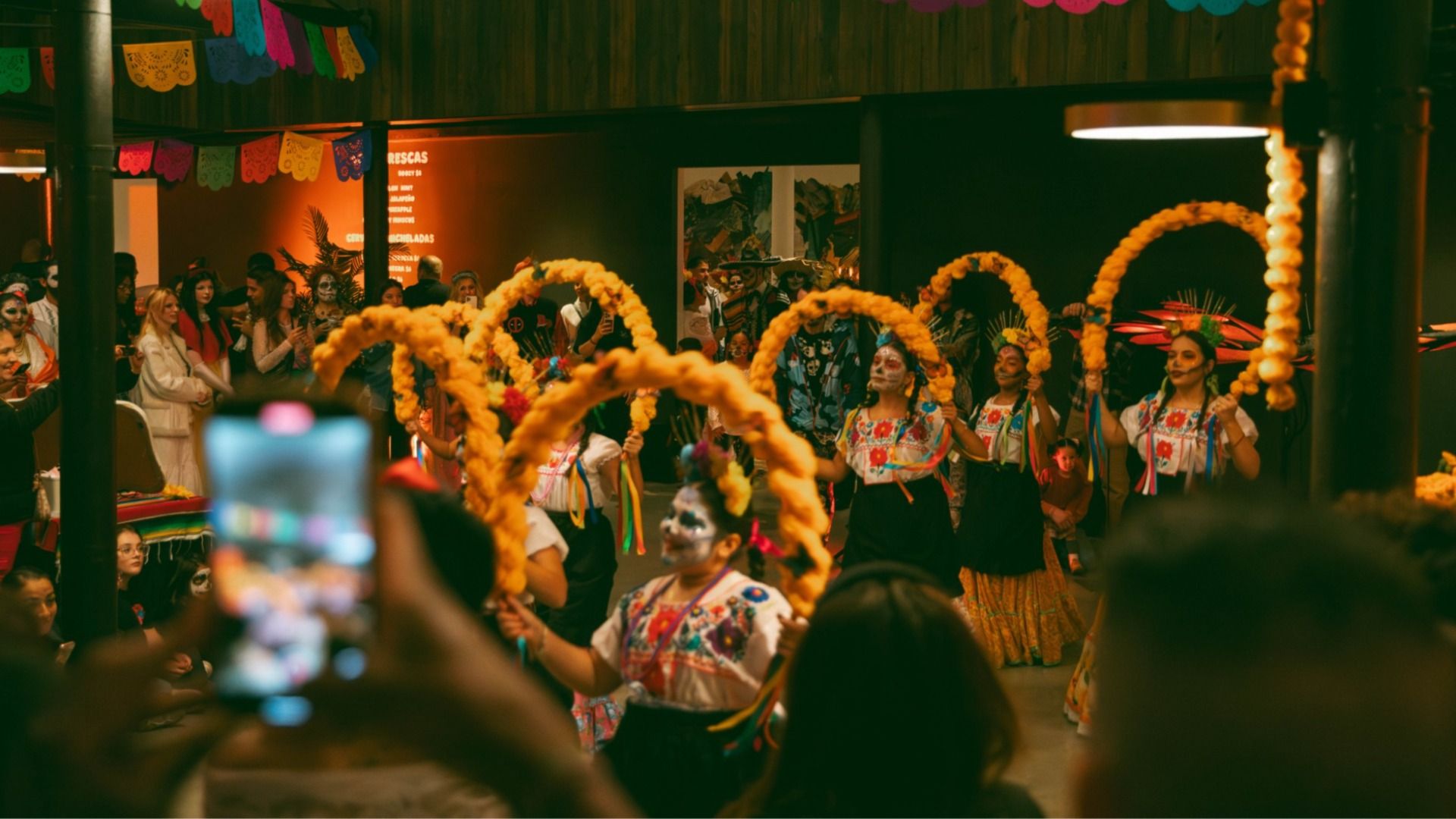Indoor festival scene: dancers in embroidered Mexican outfits perform with bright yellow flower arches while spectators film; colorful pennant banners hang overhead.