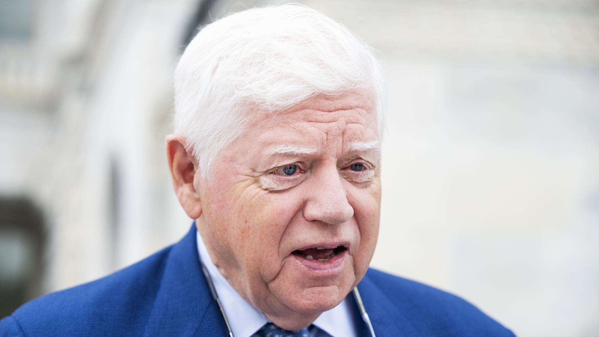 Rep. John Larson, wearing a blue suit with glasses hanging around his neck, stands outside a white marble building.
