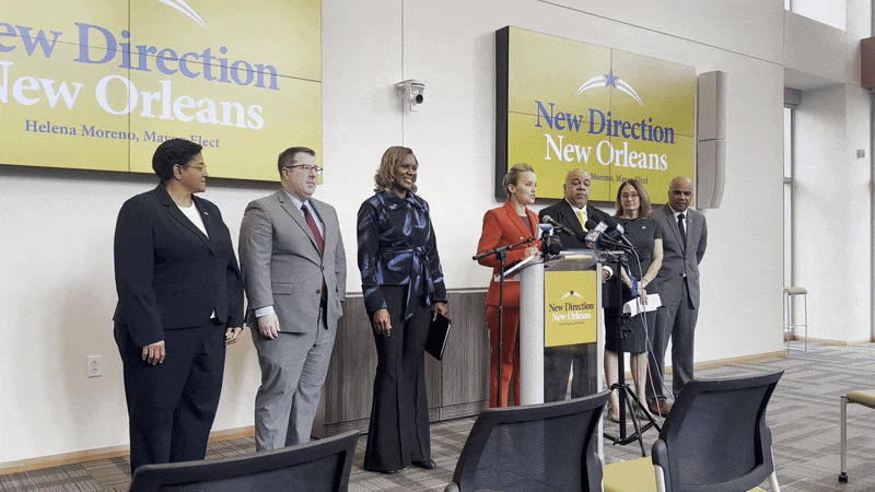 Mayor-elect Helena Moreno, standing at a podium, speaks while a line of six other people stand to either side of her.