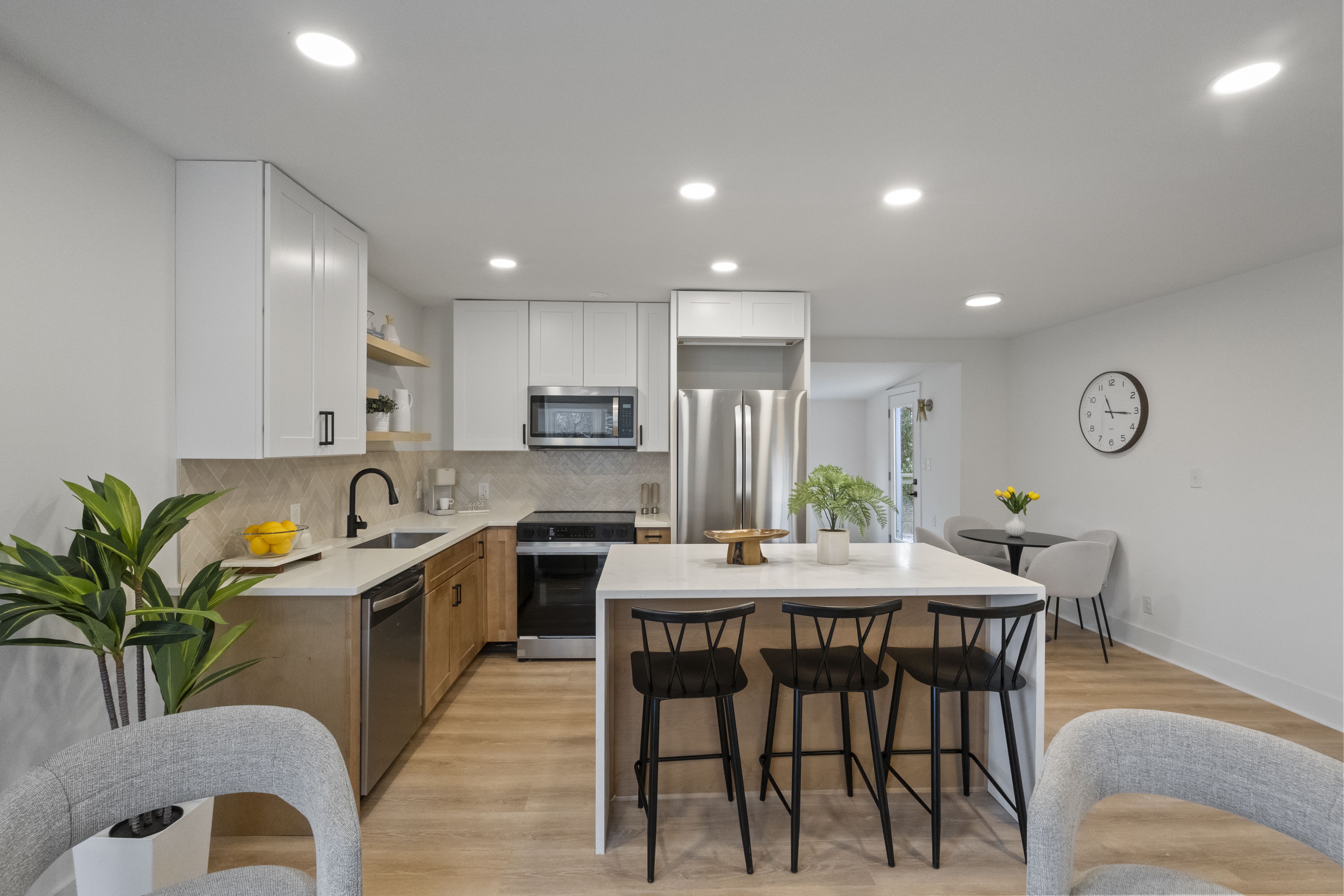 Modern kitchen with white and wood cabinets, marble island with three black stools, stainless steel appliances, a plant on the island, and a dining area with gray chairs and a clock showing 9:15.
