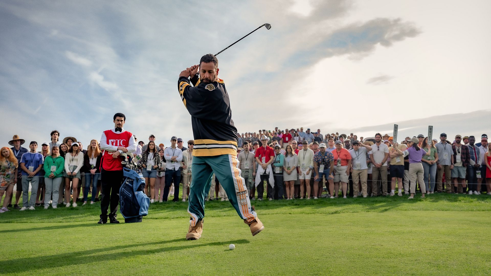 Golfer in black and yellow top with green Adidas pants prepares to swing on green golf course with large crowd watching behind under cloudy sky.