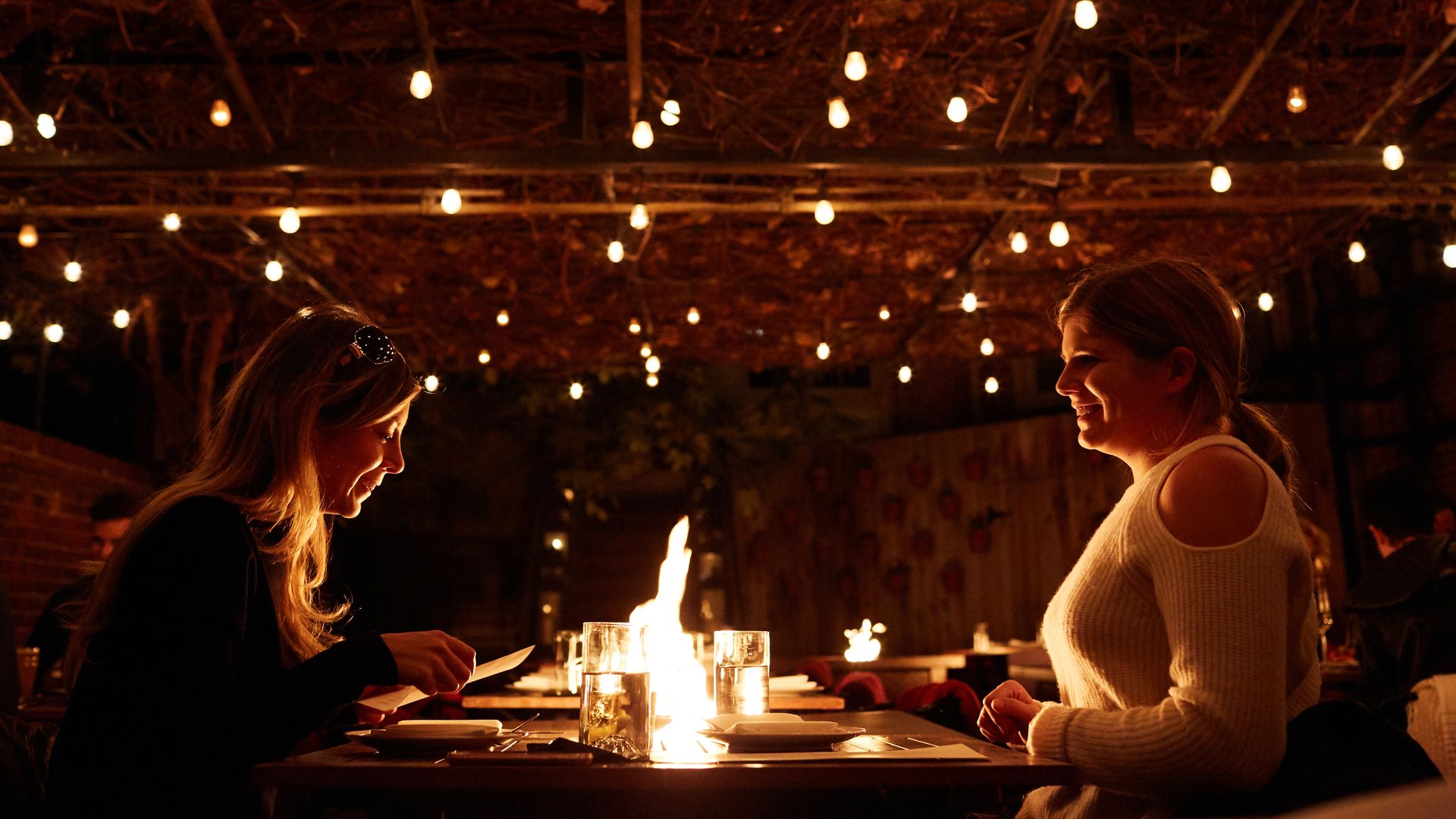 Two women sit on either sides of a fire with twinkling lights above