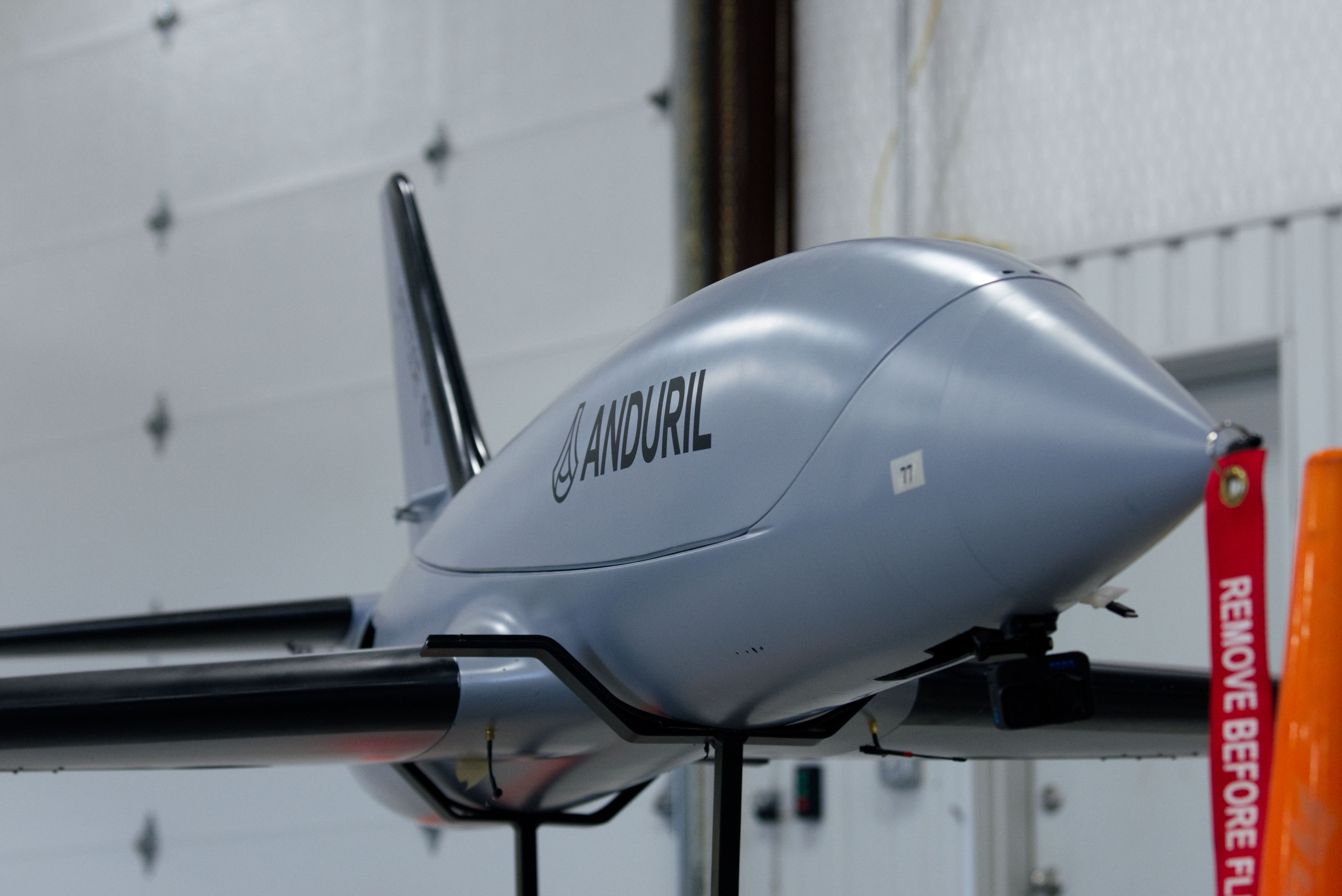 A so-called clay pigeon jet sits in a warehouse. The walls are white. The UAV is grey. A red "remove before flight" tag hangs from the nose.