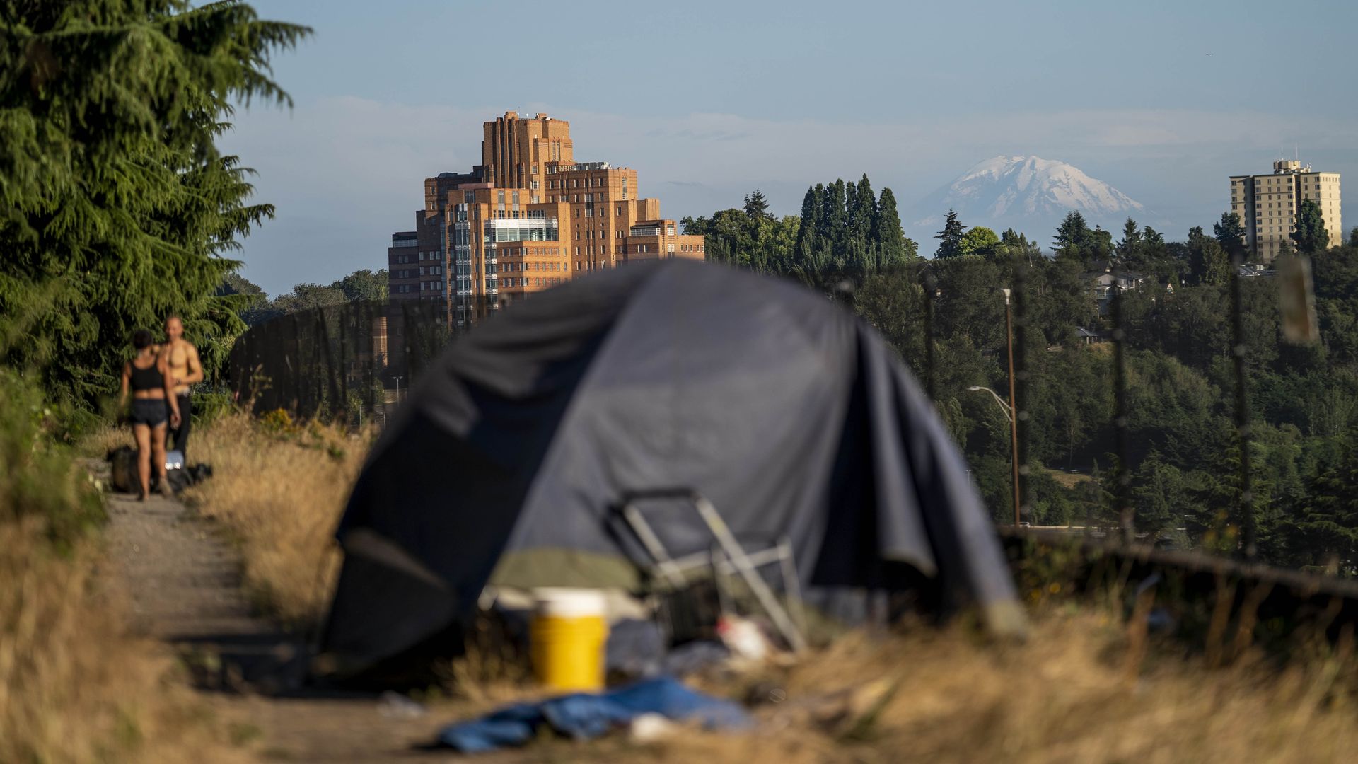 A blurred tent with belongings in dry grass along a path, two people walking, a brick building, trees, and a snow-capped mountain in the distant blue sky.
