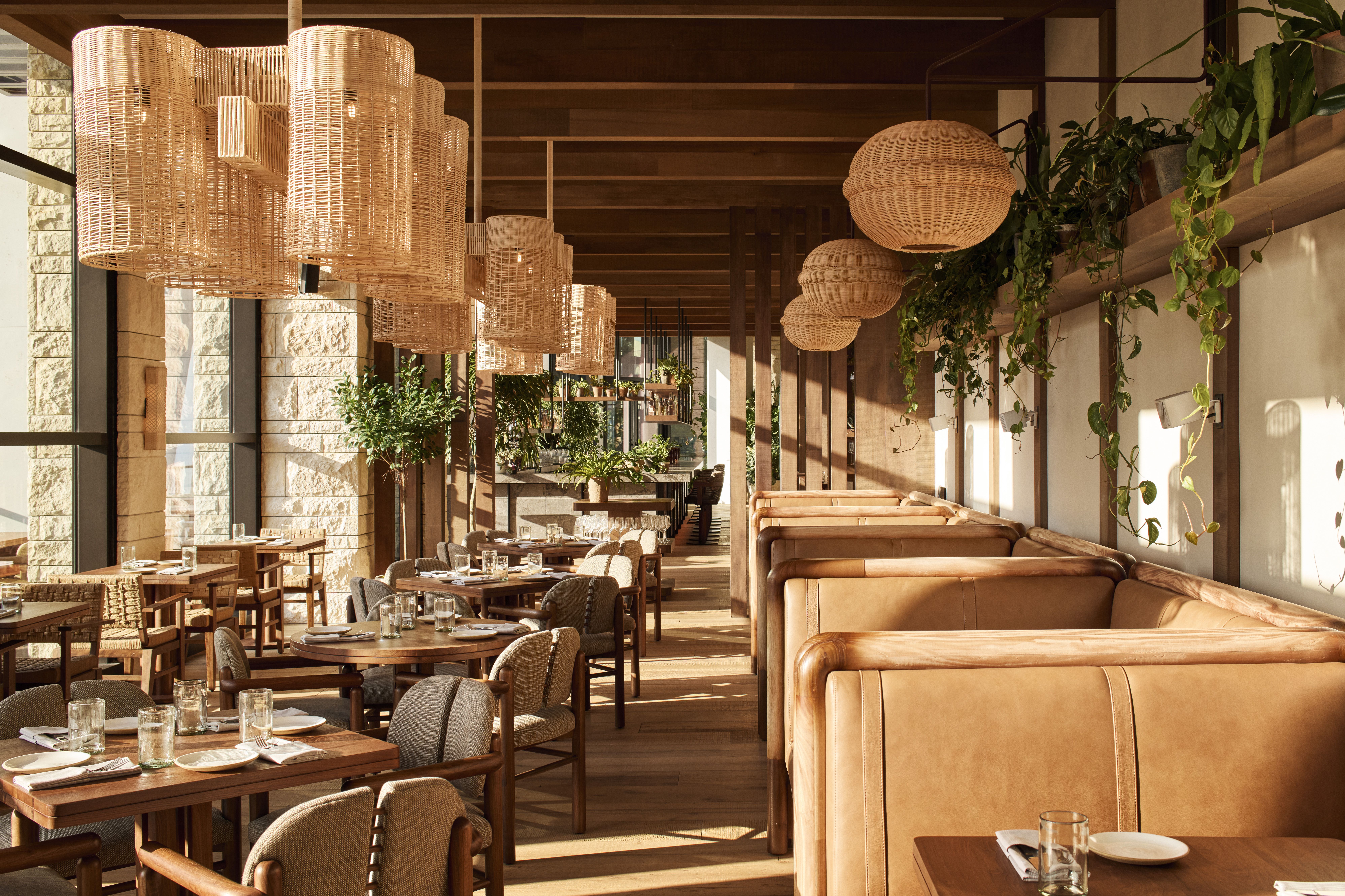 Warm restaurant interior with woven wicker pendant lights, wooden beams, stone walls, and green plants. Tables set with plates and glasses along a dining hall with tan leather booths.