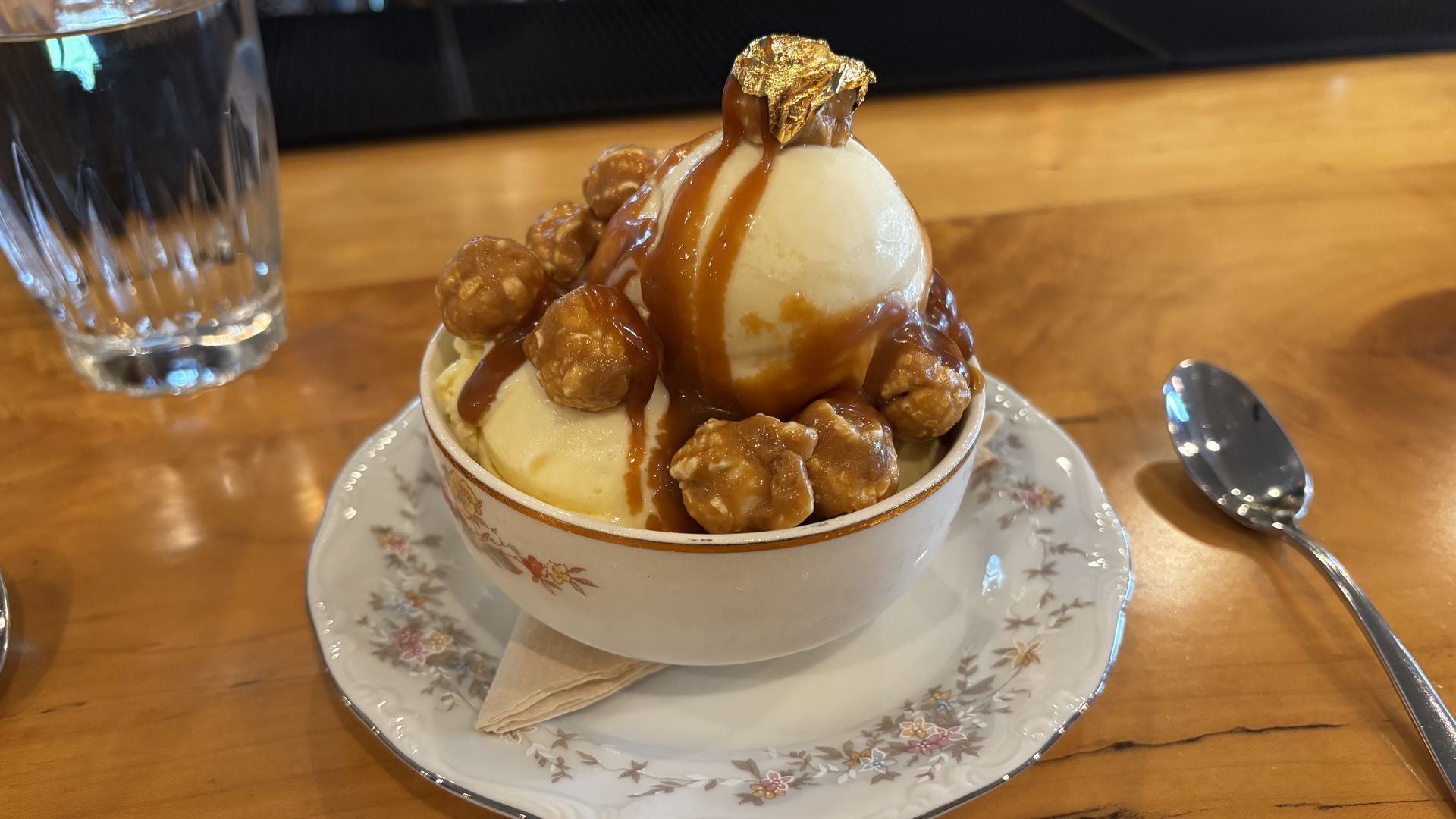 Vanilla ice cream scoop with caramel drizzle and edible gold leaf, surrounded by caramel-coated nuts in a floral teacup on a saucer, set on a wooden bar with a glass to the left.