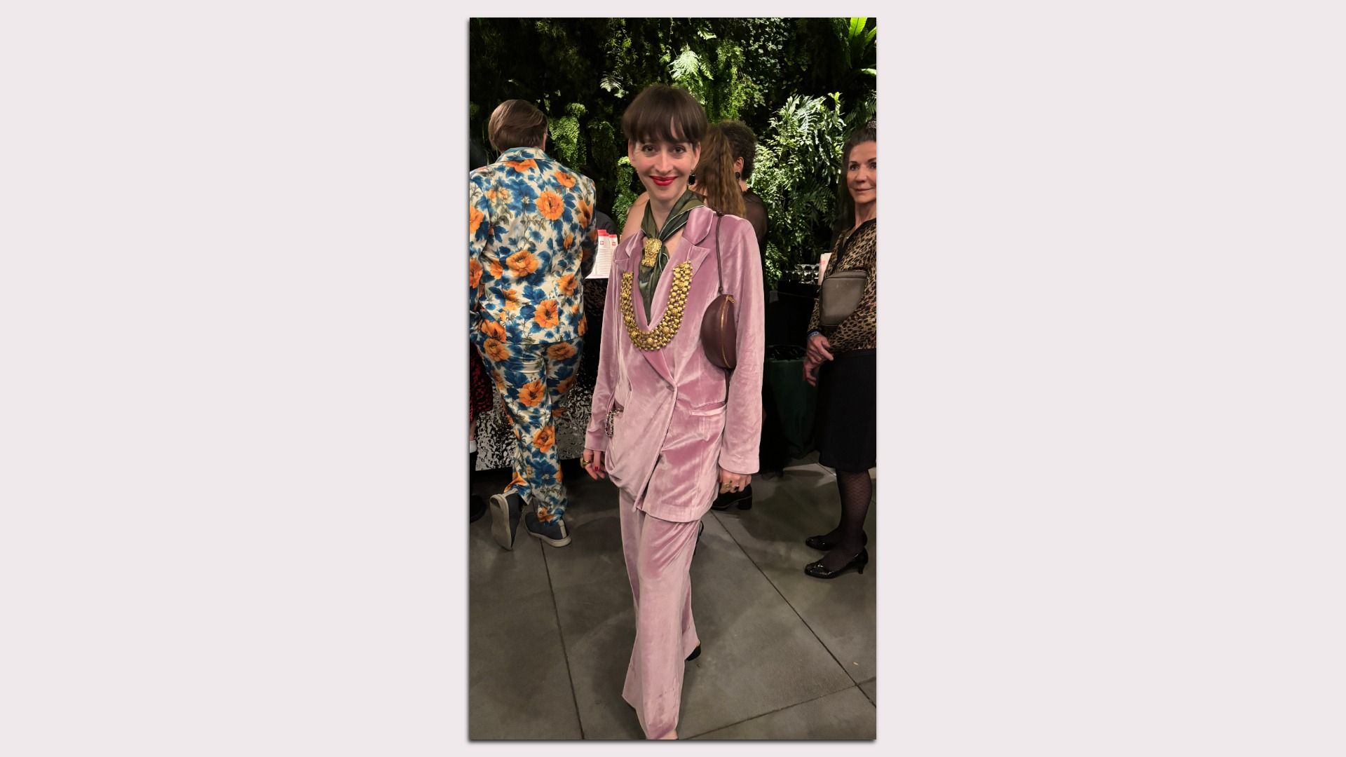 Woman in a pink velvet suit with a chunky gold necklace and green scarf posing at a fashion event; in the background are other guests in floral outfits and lush greenery.