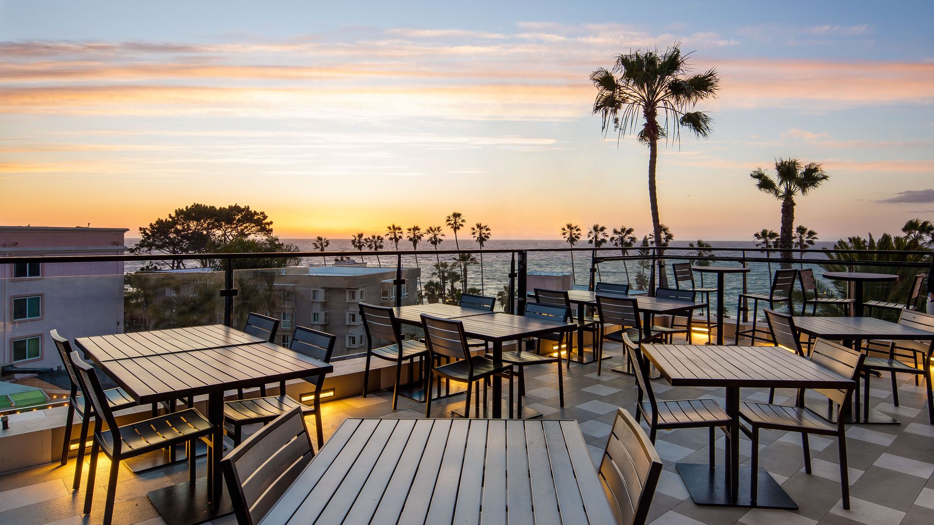 Tables at a rooftop bar with a pink and blue sunset over the ocean and palm trees in the background.