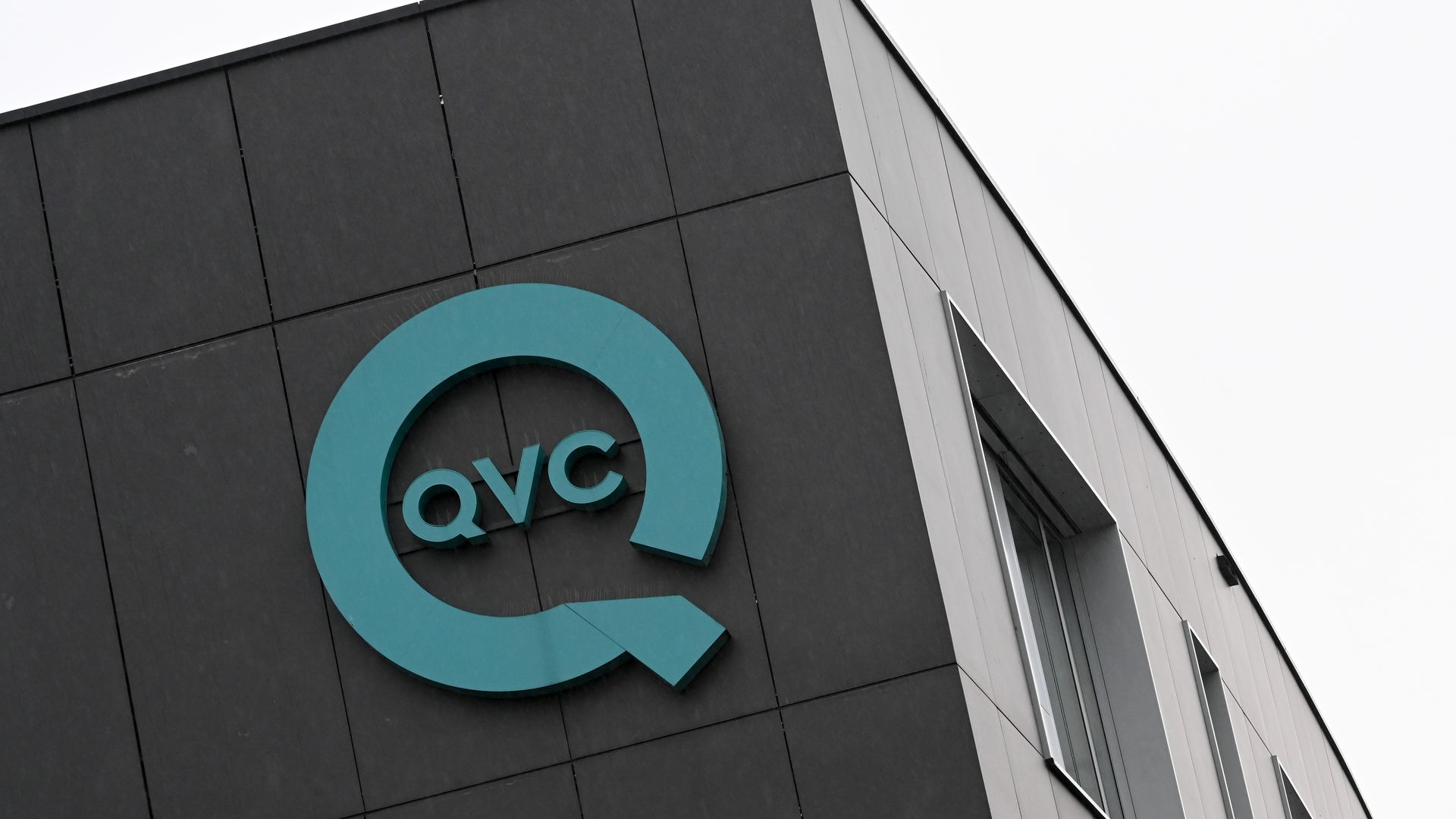 Teal QVC logo on the dark gray corner of a modern building with large windows against a white sky.