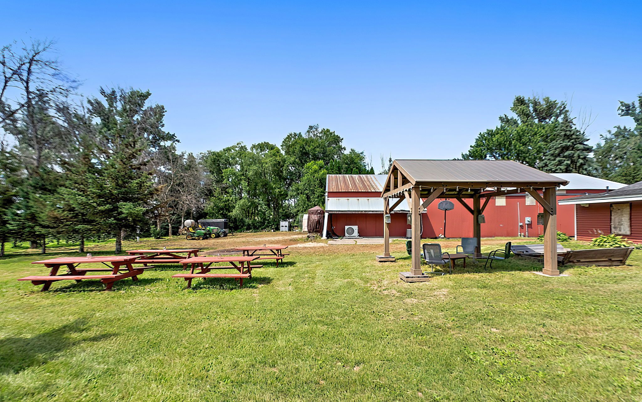 Outdoor area with red picnic tables on green grass, wooden gazebo with chairs, hammock, and red barn buildings under a clear blue sky surrounded by trees.