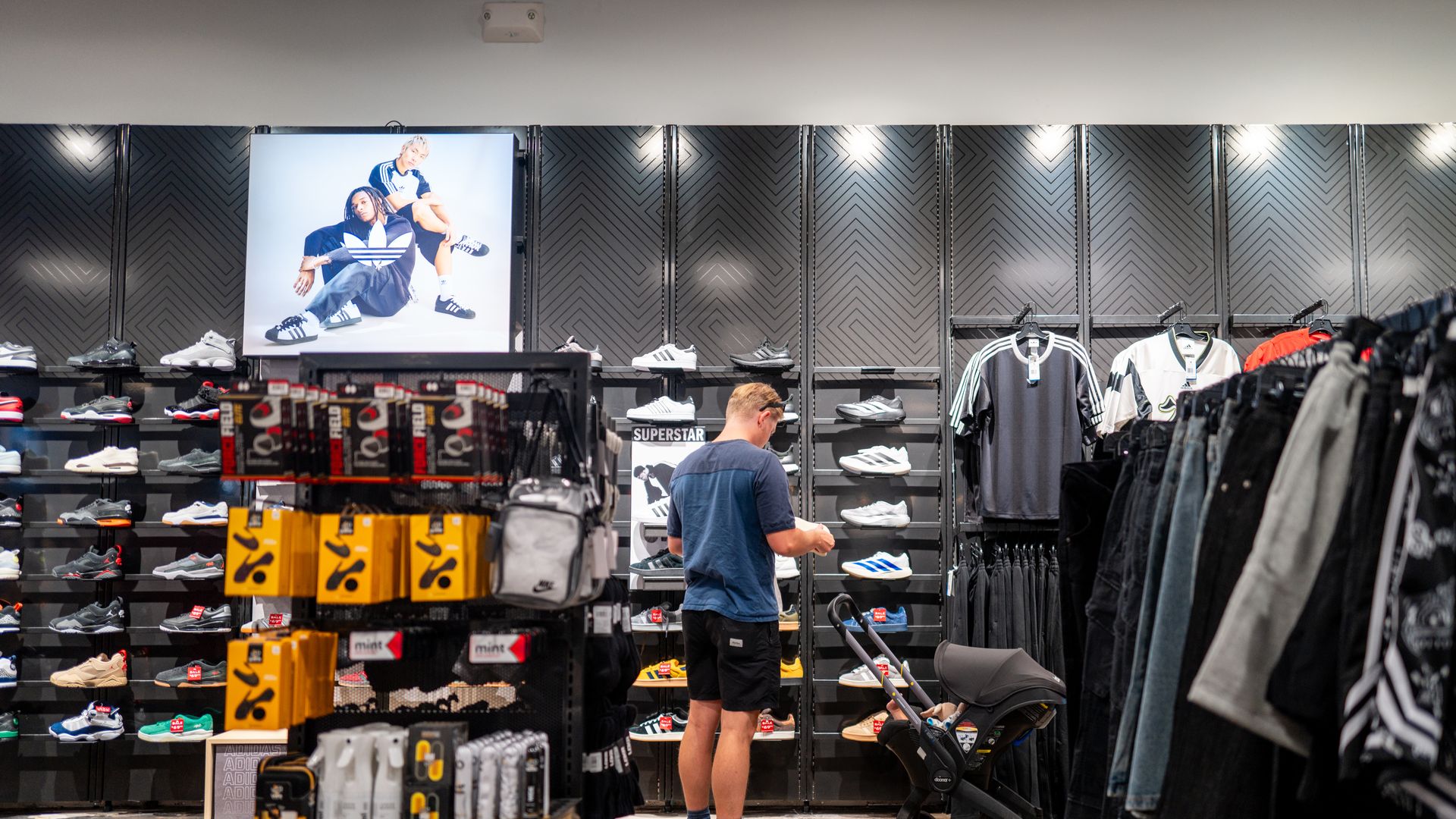 A man looking at shoes in front of a wall of sneakers at a shoe store. 