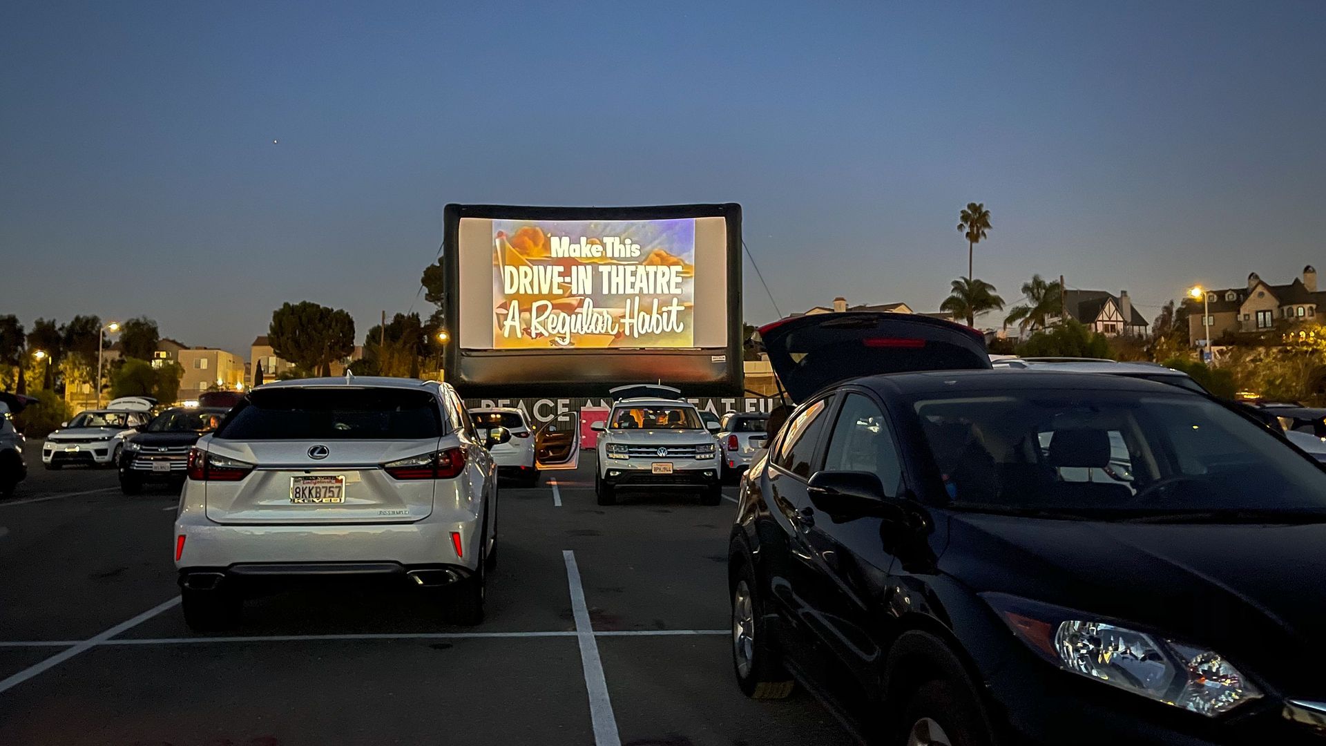 Evening at a drive-in theater with cars parked facing a large screen displaying the message, \"Make This DRIVE-IN THEATRE A Regular Habit\" under a clear blue sky.