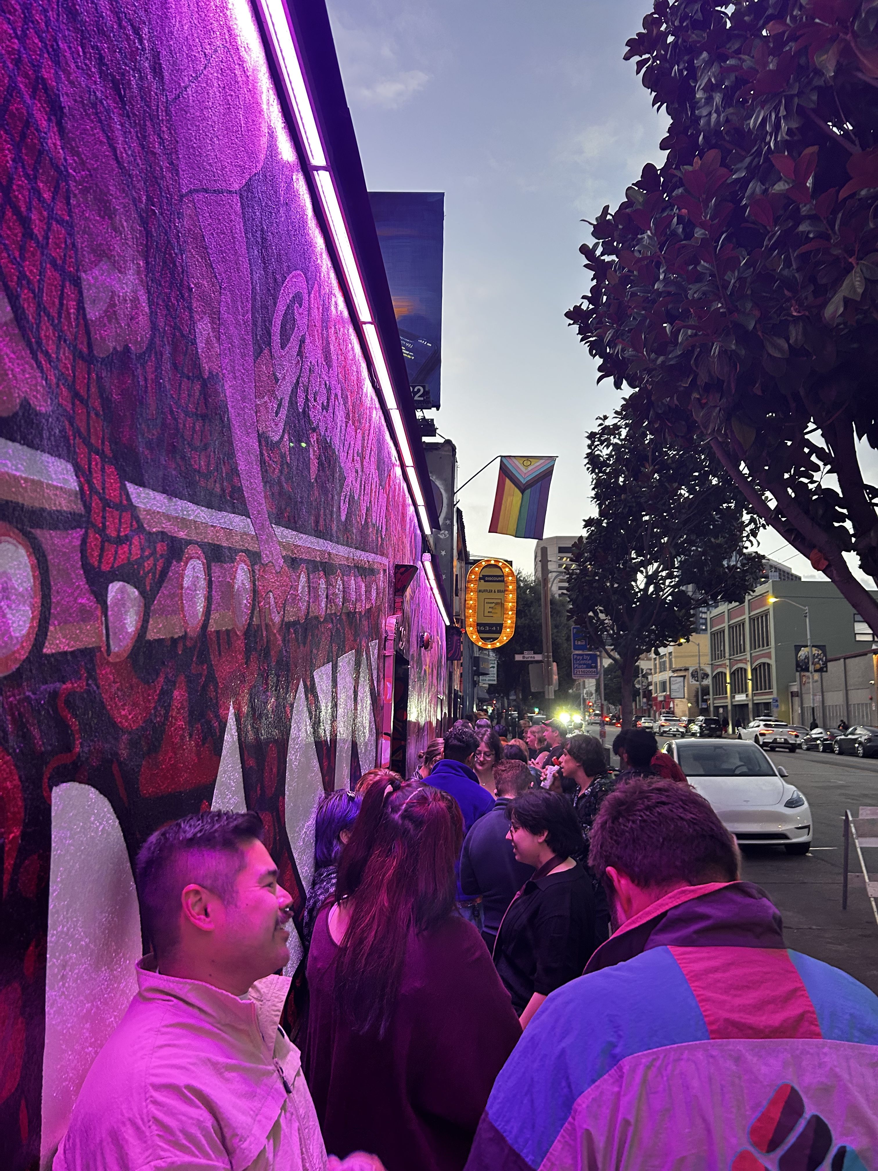 Photo of people waiting in line outside the Oasis nightclub, whose entrance is adorned with an LGBTQ flag and a giant, glittering "O"