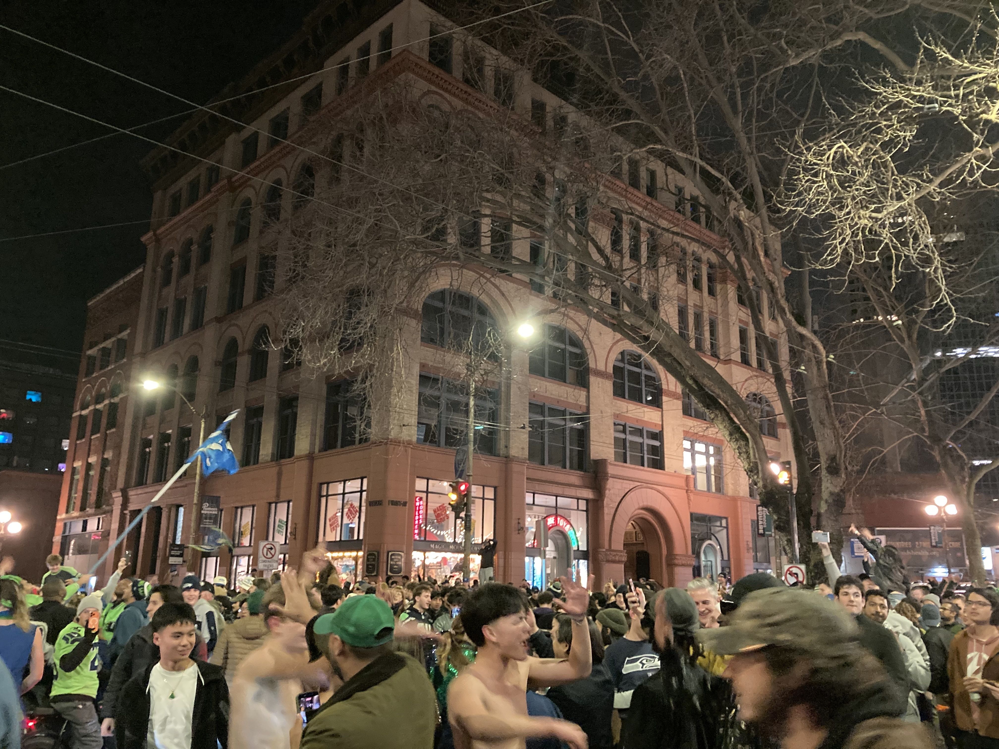 Crowd of people celebrating energetically outside a tall brick building at night, many wearing green and blue, some waving flags, with leafless trees and streetlights illuminating the scene.